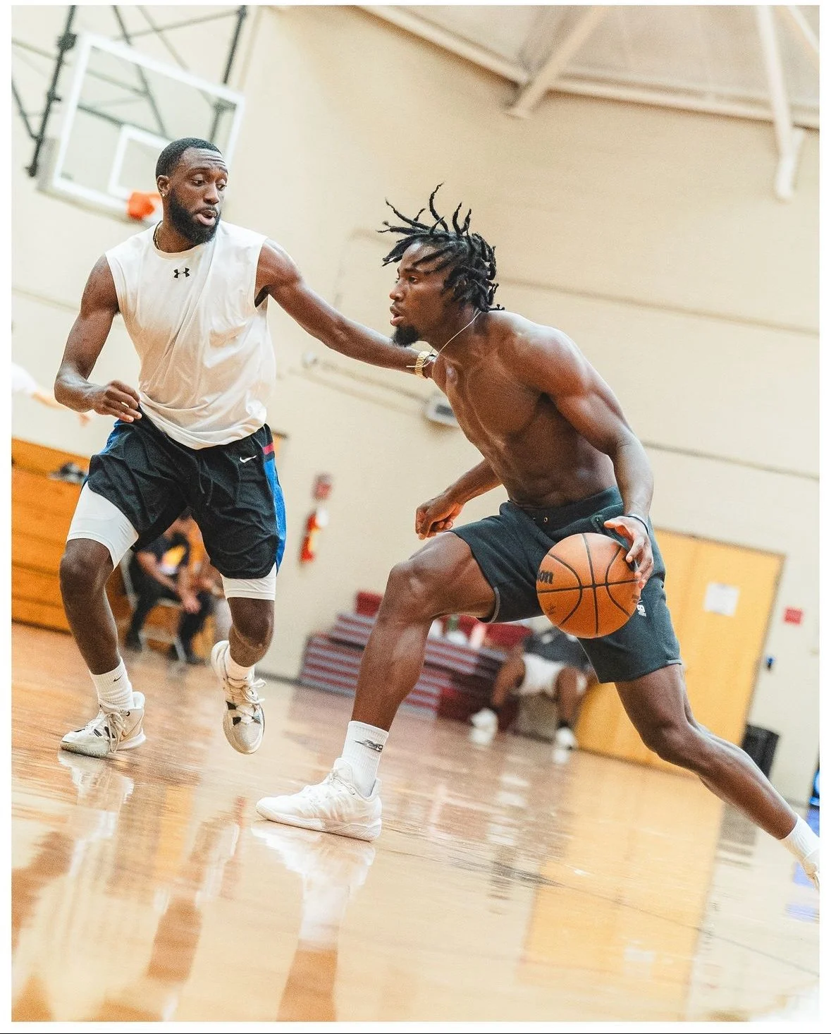Two basketball players competing for the ball on an indoor court; one is shirtless with dreadlocks, dribbling the ball, while the other is wearing a sleeveless shirt and shorts, defending.