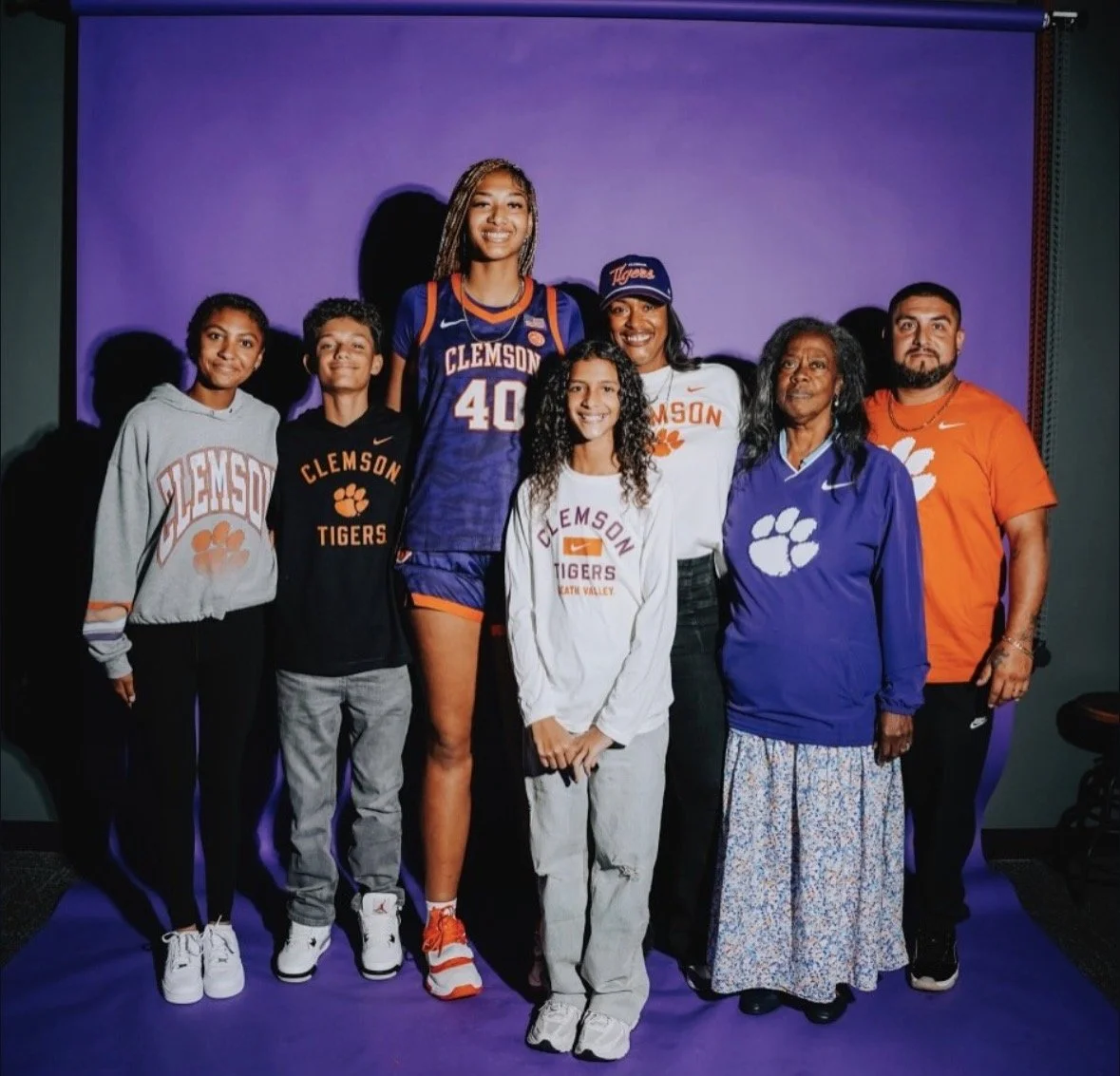 Group photo of seven people, including a female basketball player in a Clemson Tigers uniform, surrounded by family and supporters, in front of a purple backdrop.