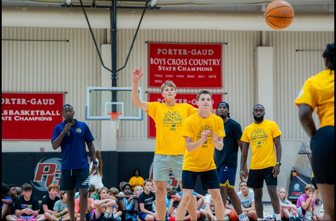 A basketball training or coaching event at a gymnasium, with a coach speaking into a microphone and four basketball players wearing yellow shirts, standing on a basketball court surrounded by children and other attendees.