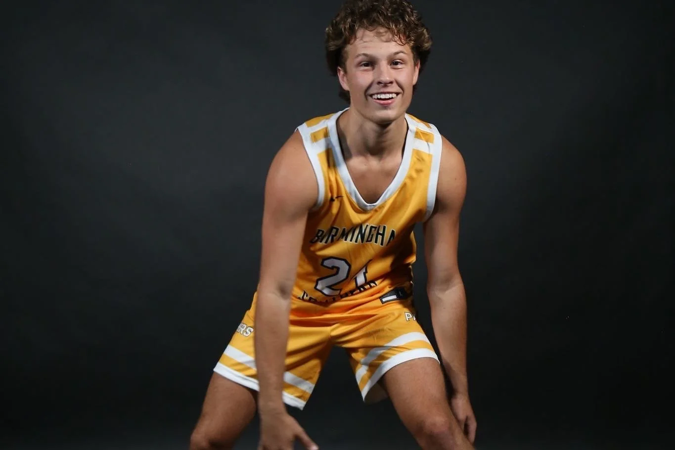 Young male basketball player wearing a yellow and white uniform with the number 21, smiling and crouching against a dark background.