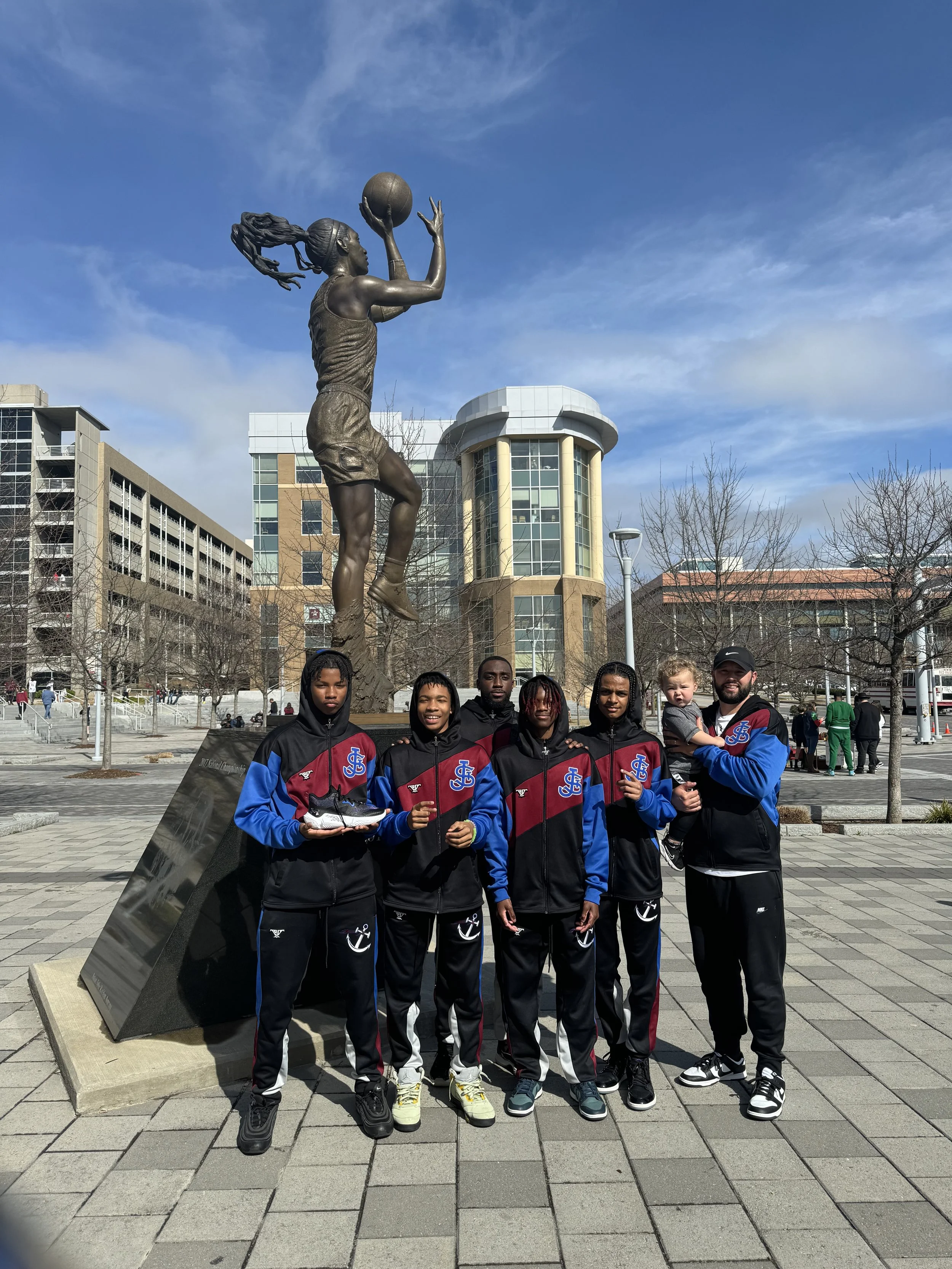 A group of six people wearing matching sports jackets are standing in front of a large statue of a female basketball player holding a basketball, located outdoors in an urban area on a sunny day.