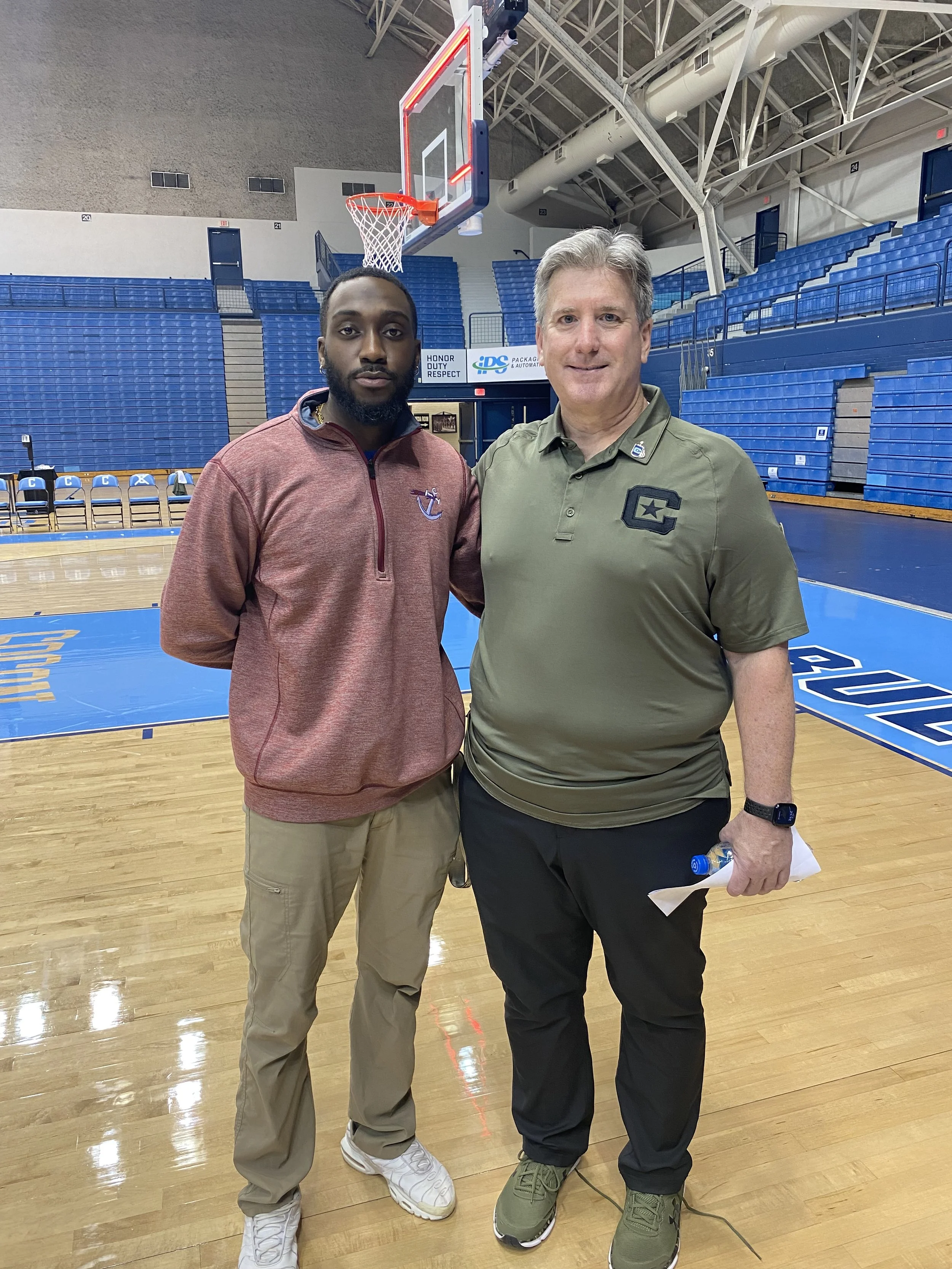 Two men standing on a basketball court, one is wearing a maroon zip-up jacket and khaki pants, the other in an olive green polo shirt and black pants, with an indoor basketball court and blue seating in the background.