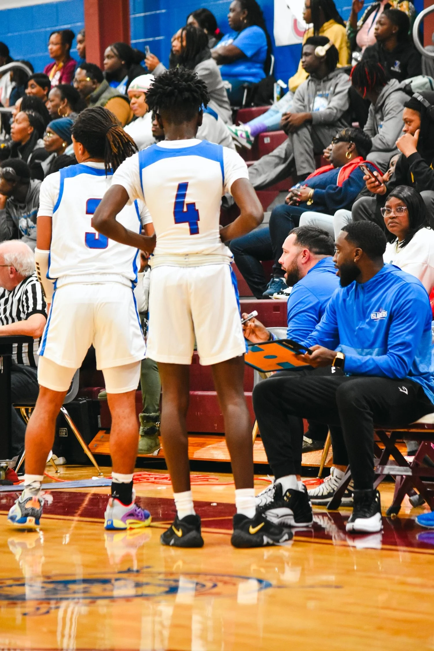 Basketball players and spectators during a game, with two players in white uniforms standing and a coach taking notes on a clipboard. Spectators are seated behind them, watching the game.