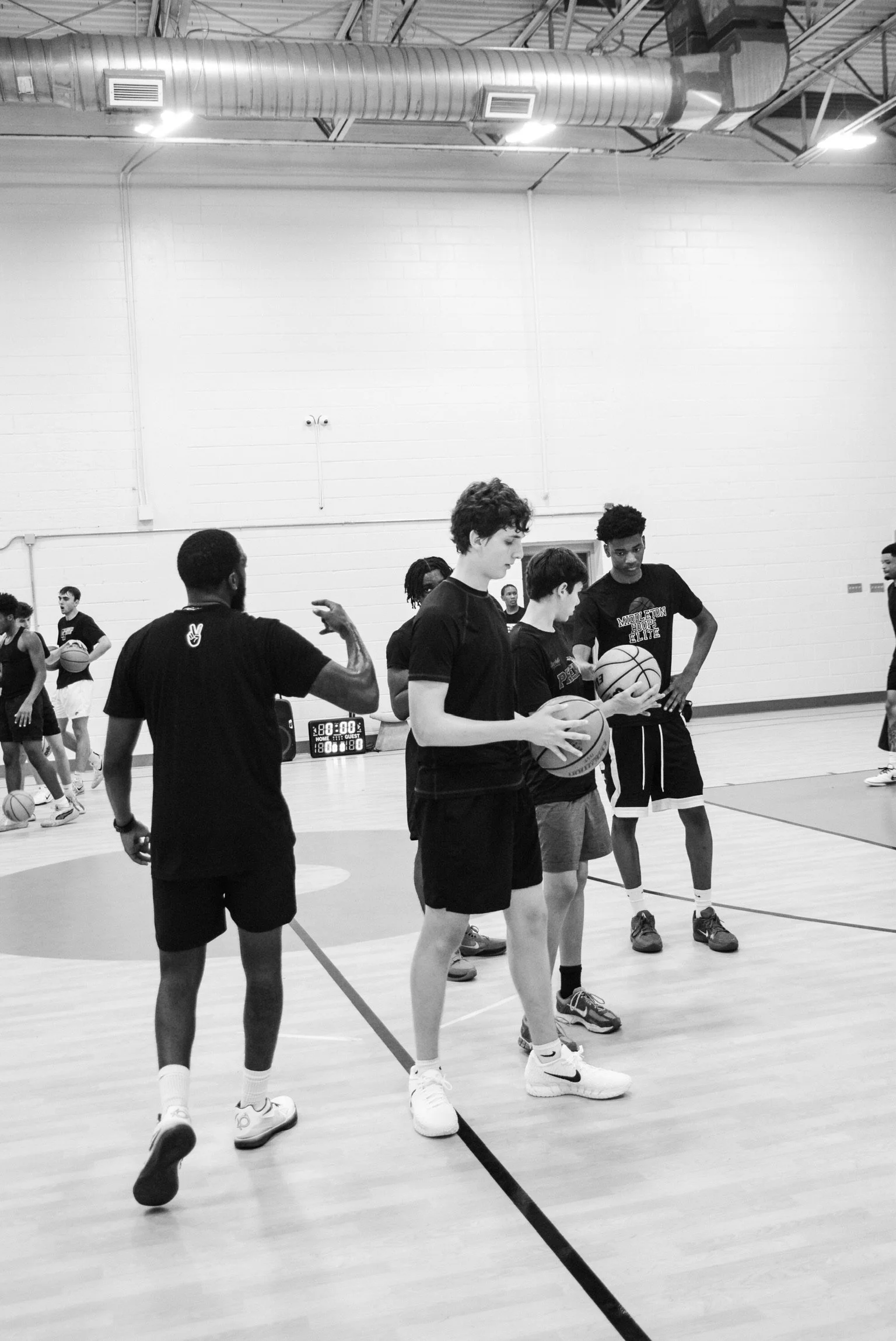 Young basketball players, some holding basketballs, are practicing on an indoor court with a coach supervising. The players appear to be in a training session or practice.