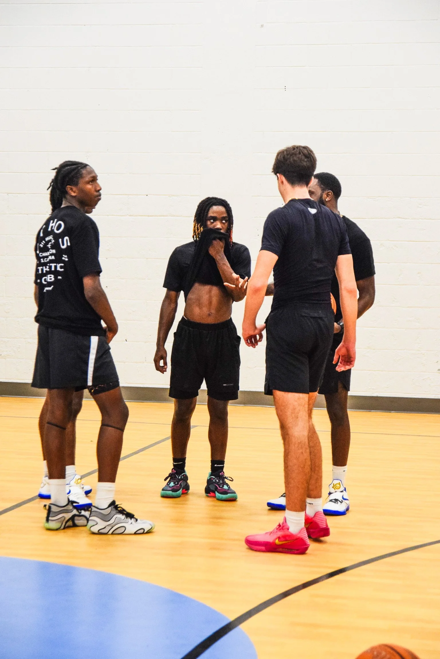 Group of young men in a gymnasium, standing in a circle, possibly having a discussion or strategizing during a basketball game or practice. One is lifting his shirt.