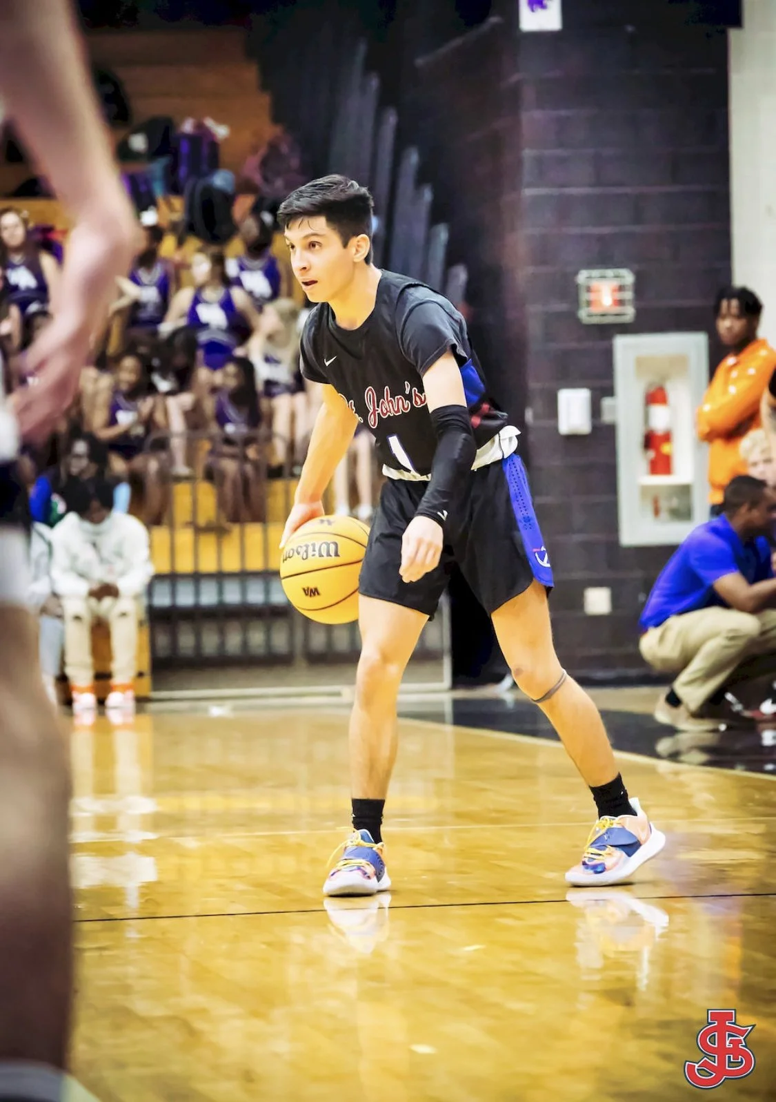 A young man playing basketball on an indoor court, wearing a black jersey with red and white lettering, black shorts, and colorful sneakers, with an audience watching in the background.