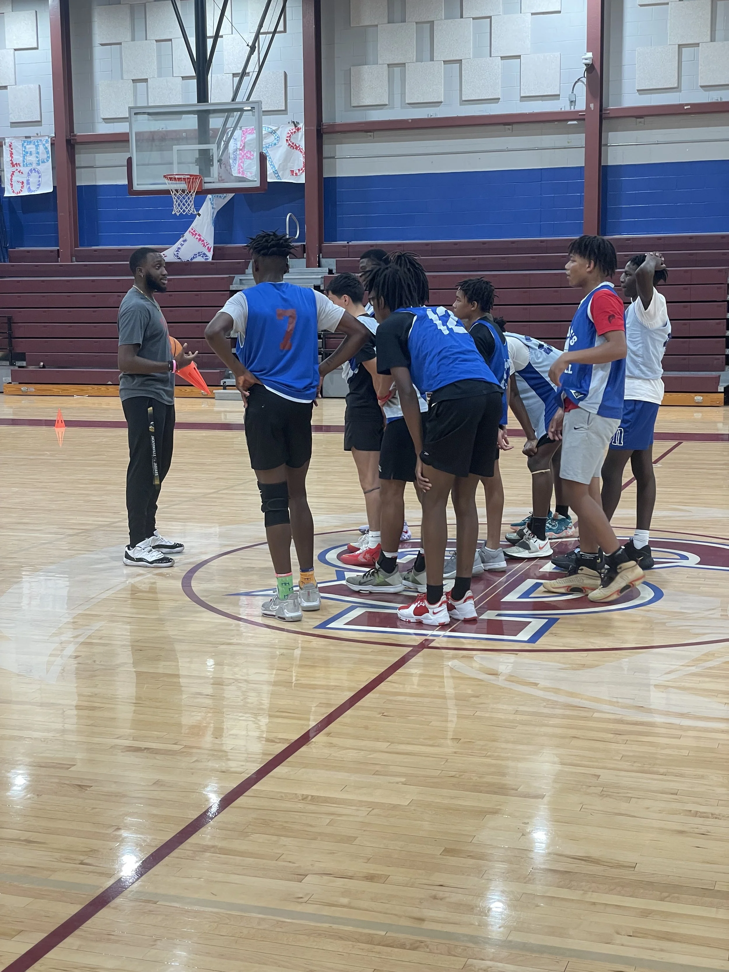 A basketball coach giving instructions to a team during a practice session in an indoor gymnasium.