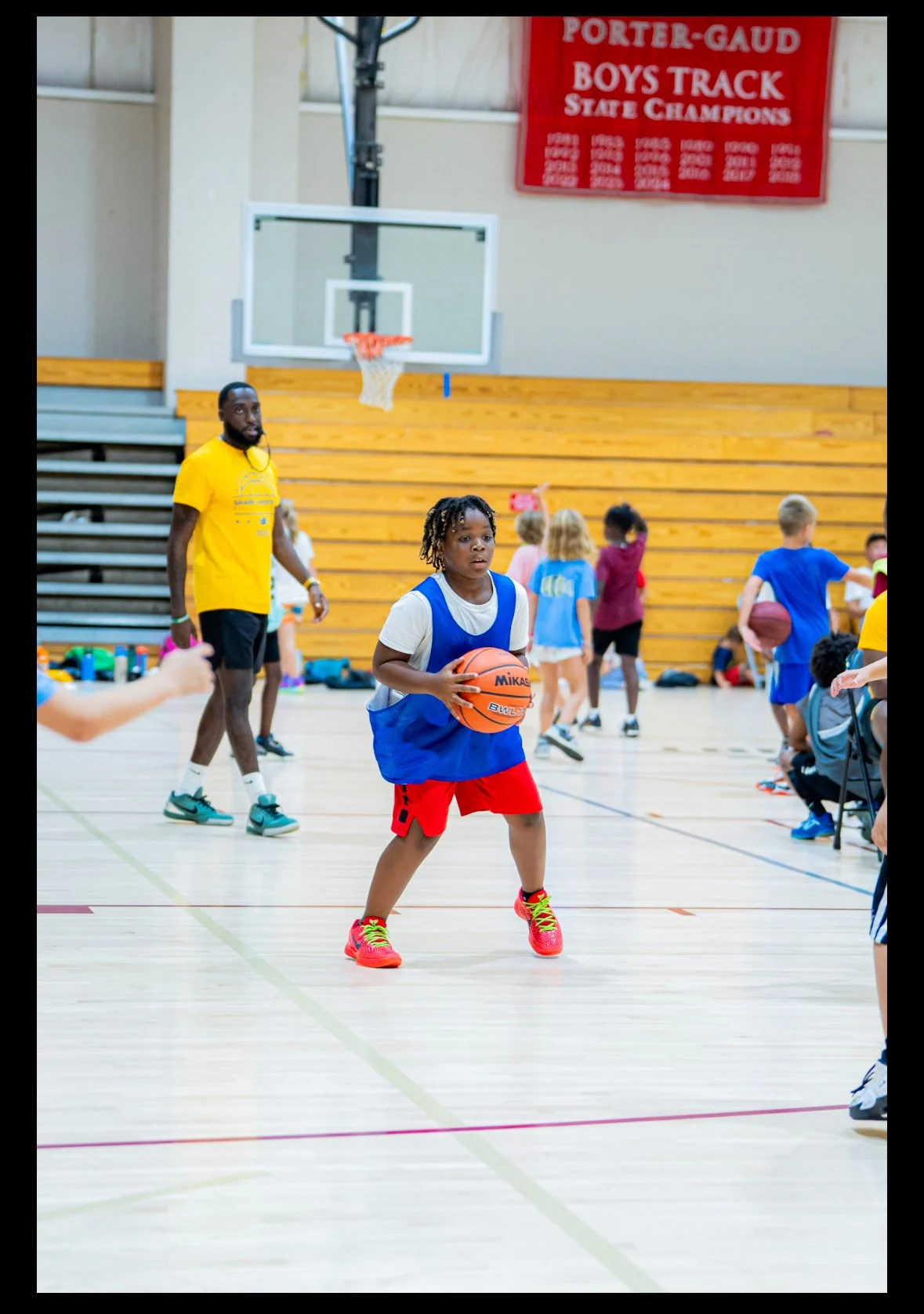 A young girl in a blue jersey and red shorts holding a basketball on an indoor basketball court, with children and a coach in the background during a basketball practice or clinic.