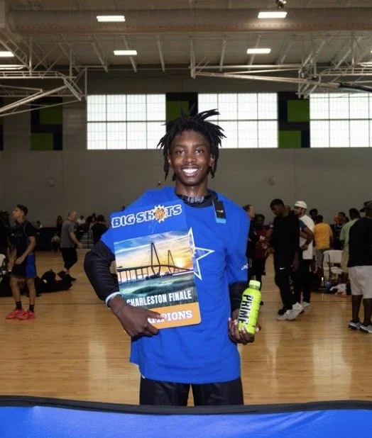 Smiling young man with dreadlocks wearing a blue sports jersey, holding a championship certificate and a green water bottle, standing in an indoor gymnasium filled with people.