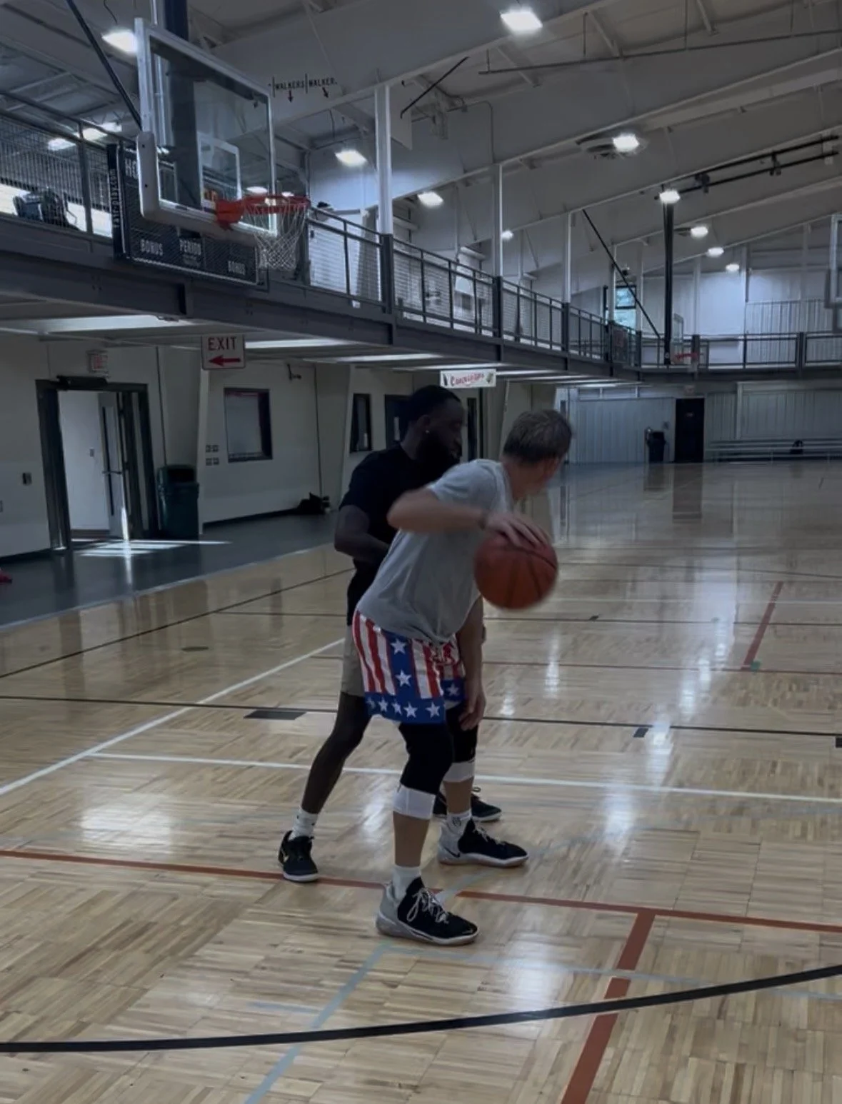 Two basketball players practicing on an indoor basketball court, one holding the ball while the other guards closely.