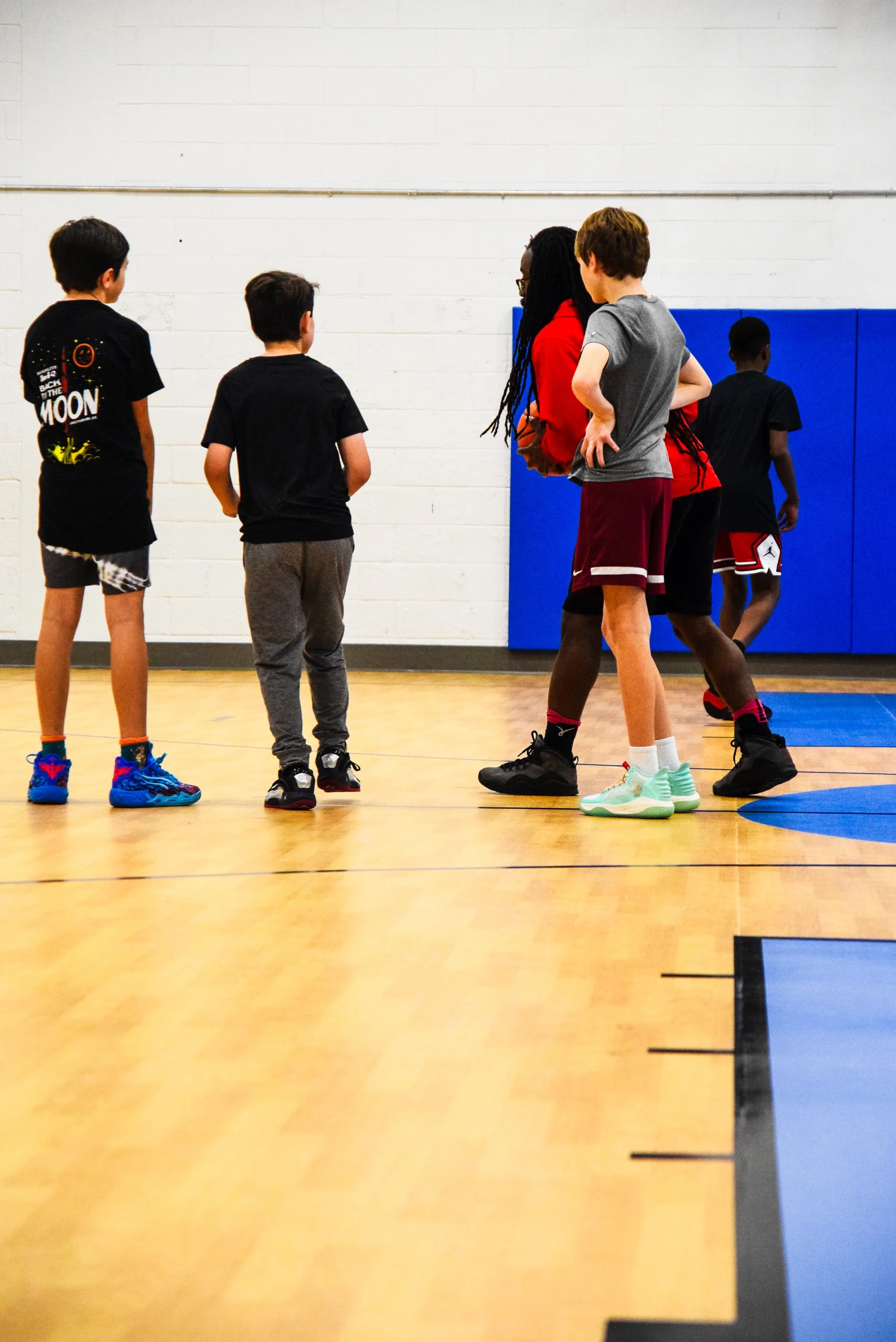 A group of five young children in a gymnasium, with some of them engaging in basketball practice. They are standing on a wooden court with a white brick wall and blue padded mat in the background.