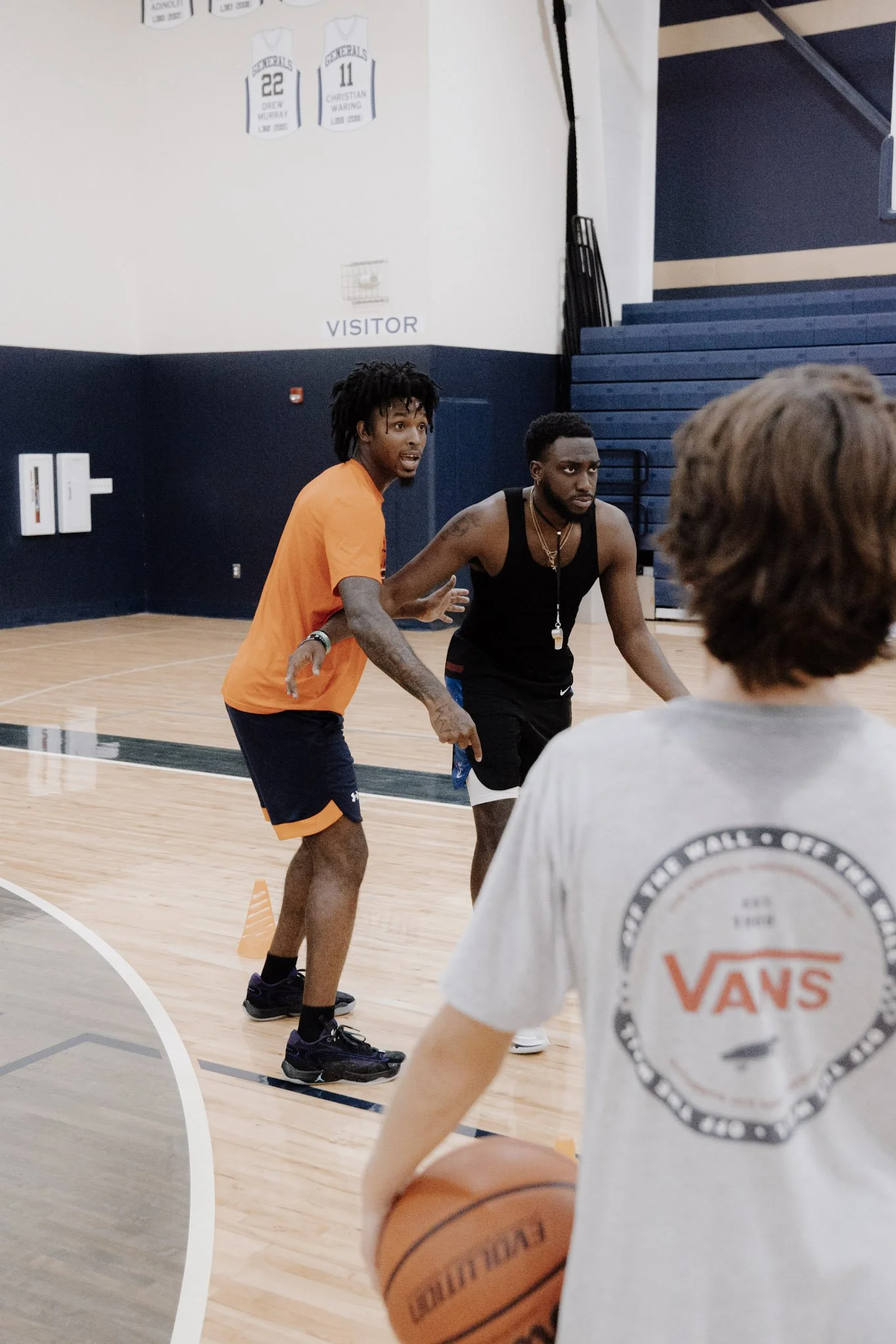 Three individuals on a basketball court in a gym, with two men coaching or instructing a young boy holding a basketball. The court has light-colored wood flooring and there are wall banners and stairs in the background.