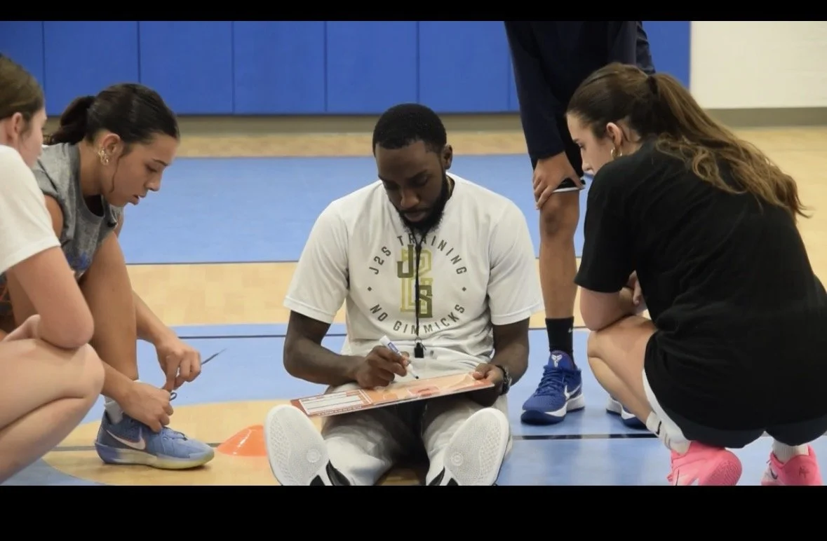 A coach seated on the gym floor with four female basketball players gathered around him, writing on a clipboard during a team practice or strategy session.