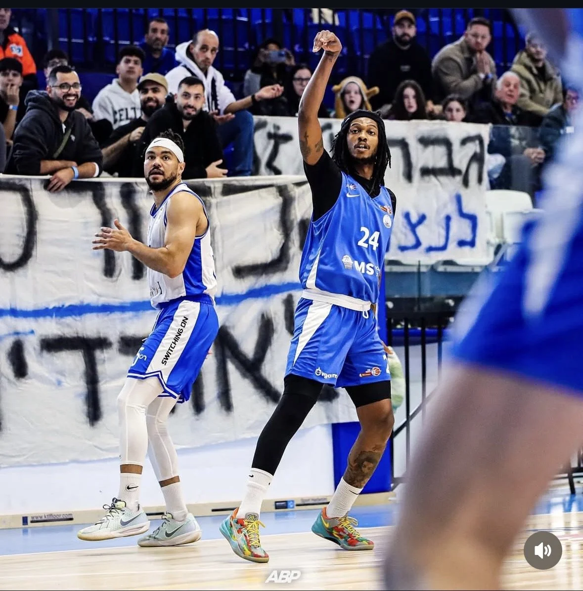 A basketball player in a blue uniform with the number 24 points towards the ceiling, with his arm raised, while another player in a white and blue uniform claps nearby. The game is taking place in an indoor basketball court, with spectators watching 