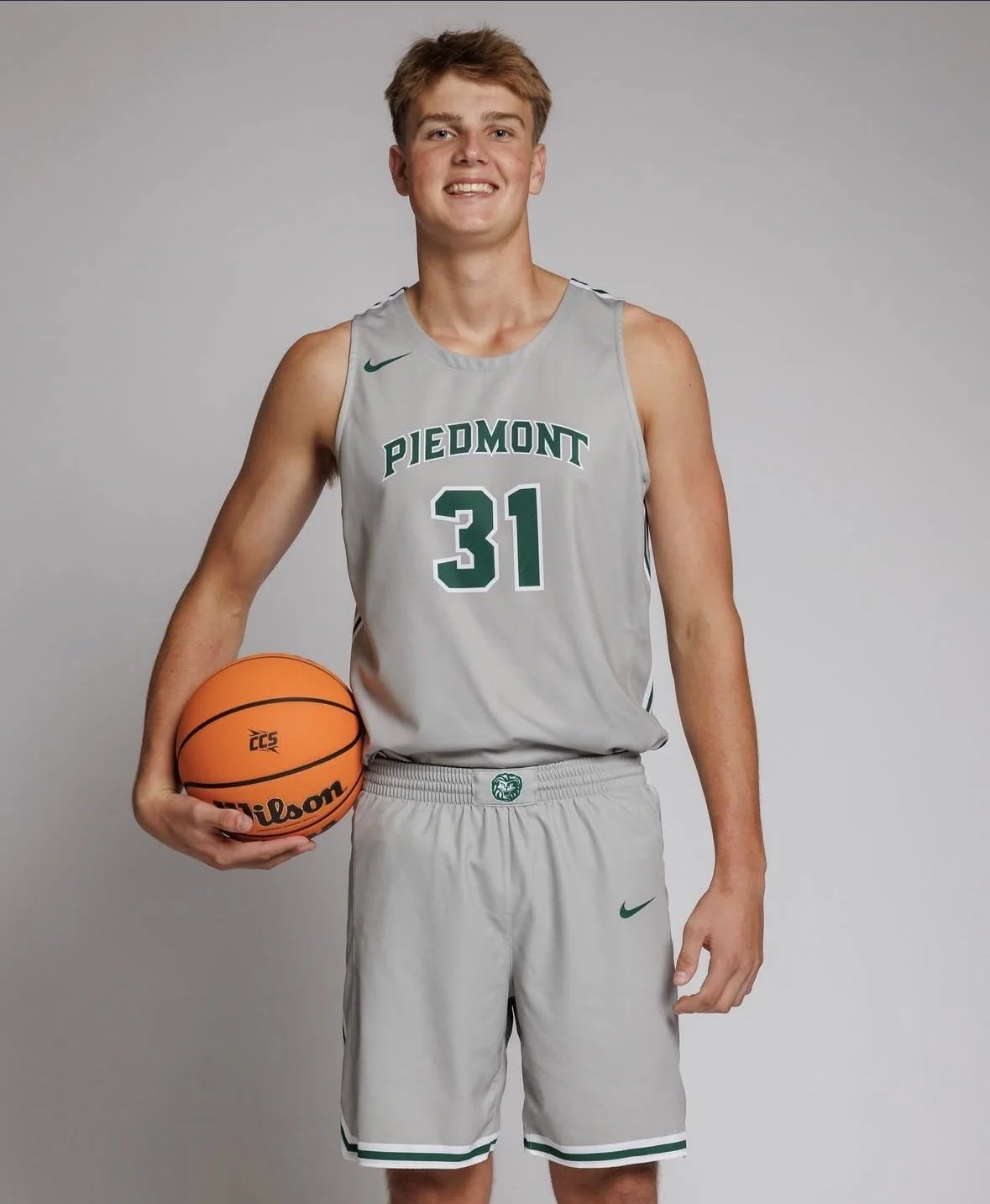 Young male basketball player in gray uniform holding an orange Wilson basketball, smiling, standing against a plain background.