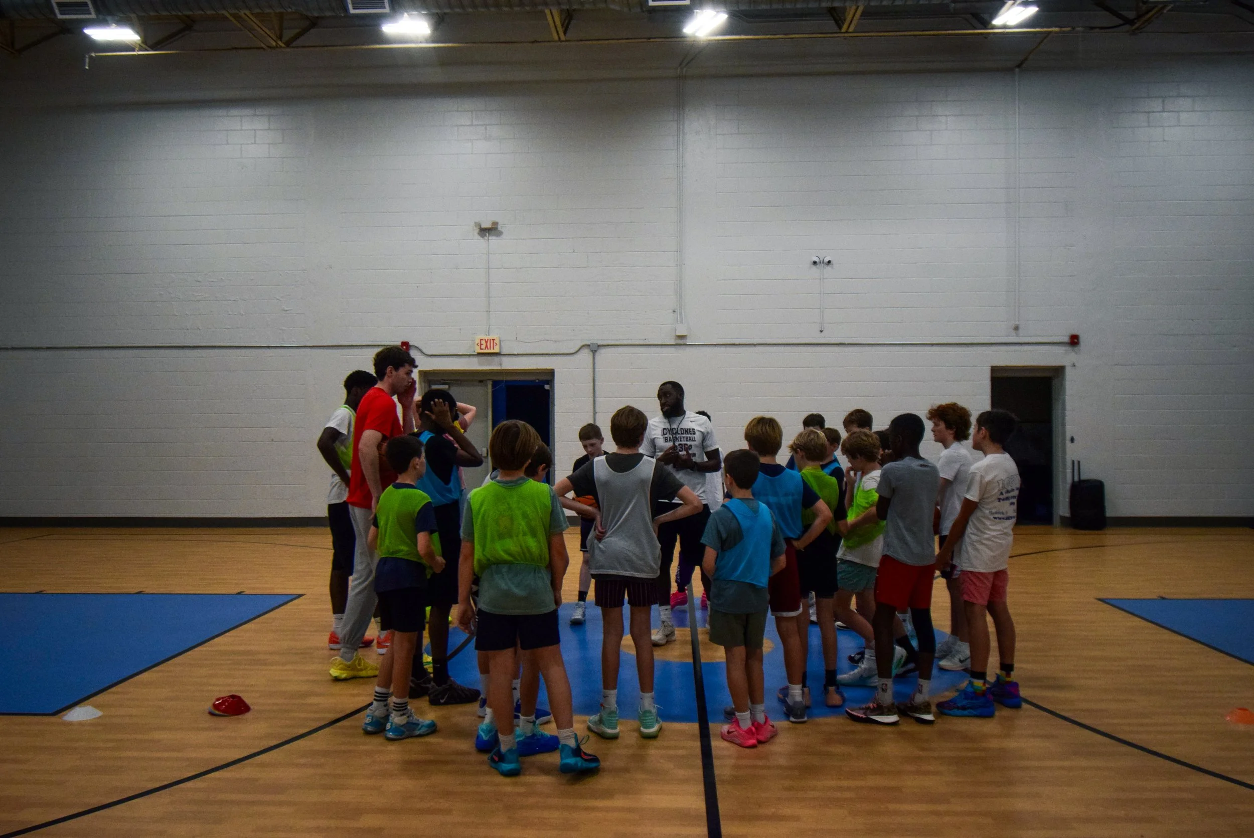 Group of children and coaches gathered in a gym for a basketball practice or instruction, with children wearing colored pinnies.