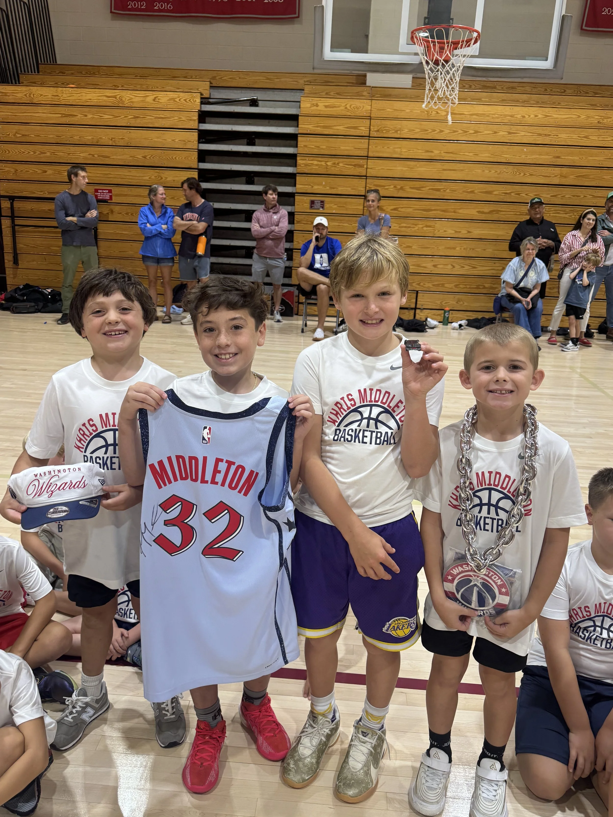 Children in basketball uniforms and t-shirts celebrating on a gymnasium court after a game, some holding jerseys, medals, and a hat, with spectators behind them.