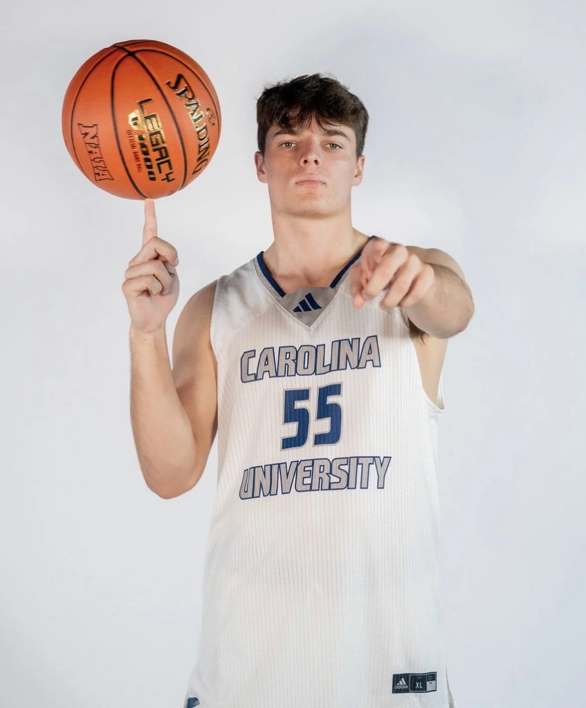 Young male basketball player in a white Carolina University jersey spinning a basketball on his index finger while pointing at the camera.