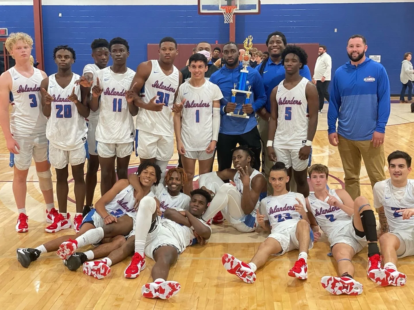 A group of basketball players and coaches celebrating with a trophy on an indoor basketball court. The players are wearing white and blue uniforms with 'Islanders' written on them, some are standing, while others are sitting or lying on the floor, al