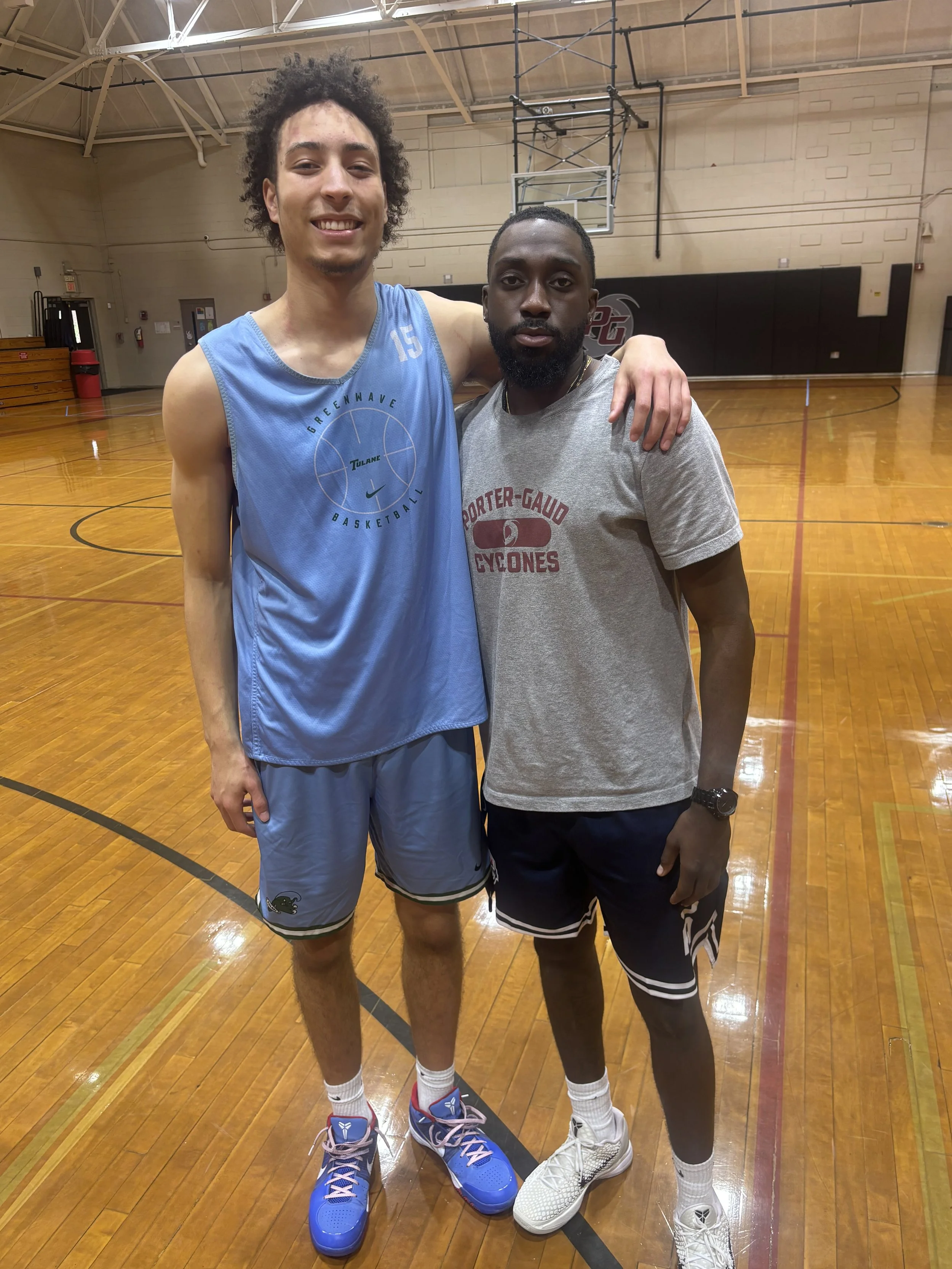 Two men standing together on a basketball court; one wearing a blue sleeveless jersey and shorts, the other in a gray t-shirt and black shorts, with their arms around each other's shoulders.