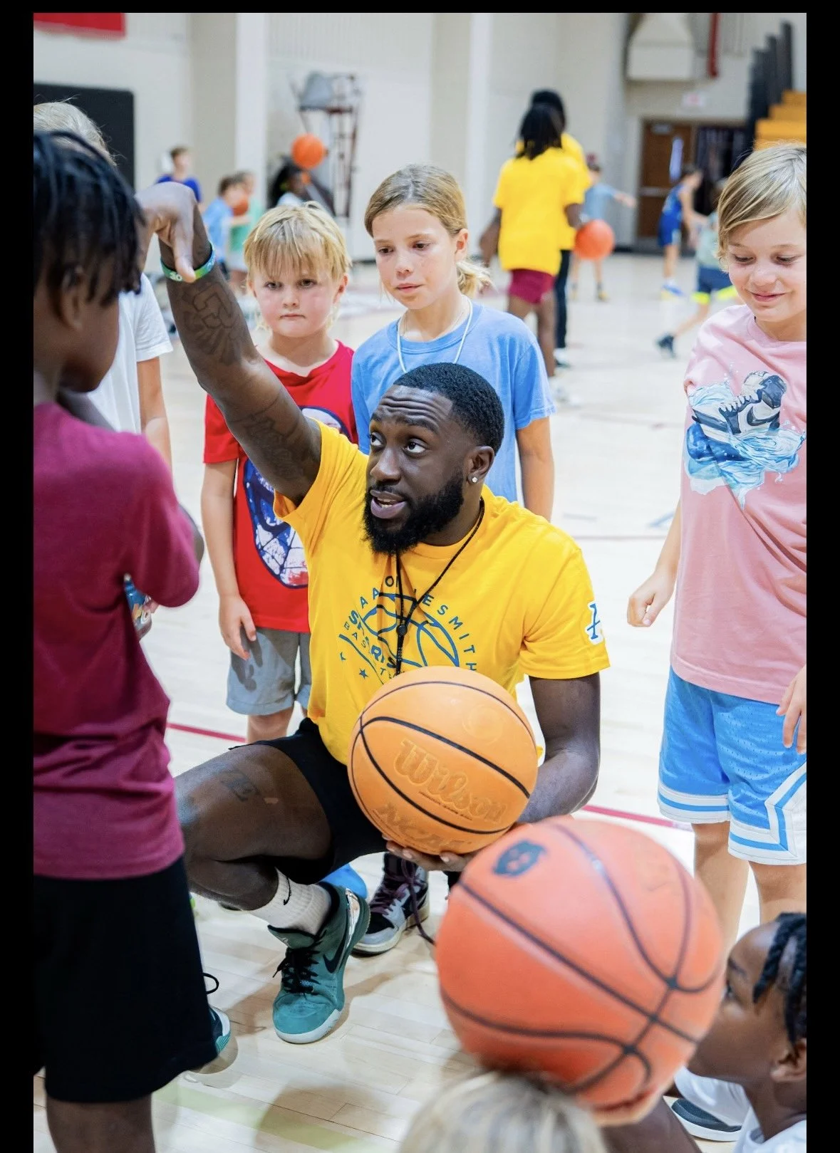 A basketball coach in a yellow shirt kneels on the gym floor, holding a basketball, instructing children during a youth basketball clinic in a gymnasium.
