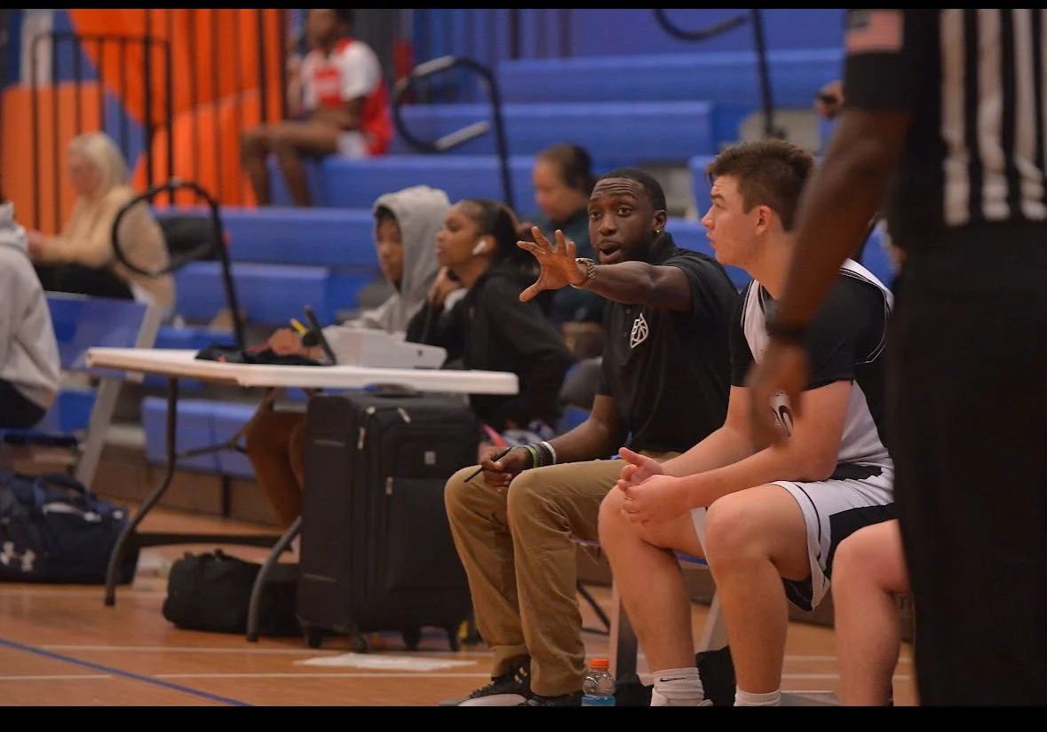 A man coaching a basketball game sitting on a bench, gesturing with his hand, wearing a black shirt, with a young basketball player in a black and white uniform sitting next to him. Several spectators are seated on blue bleachers in the background.