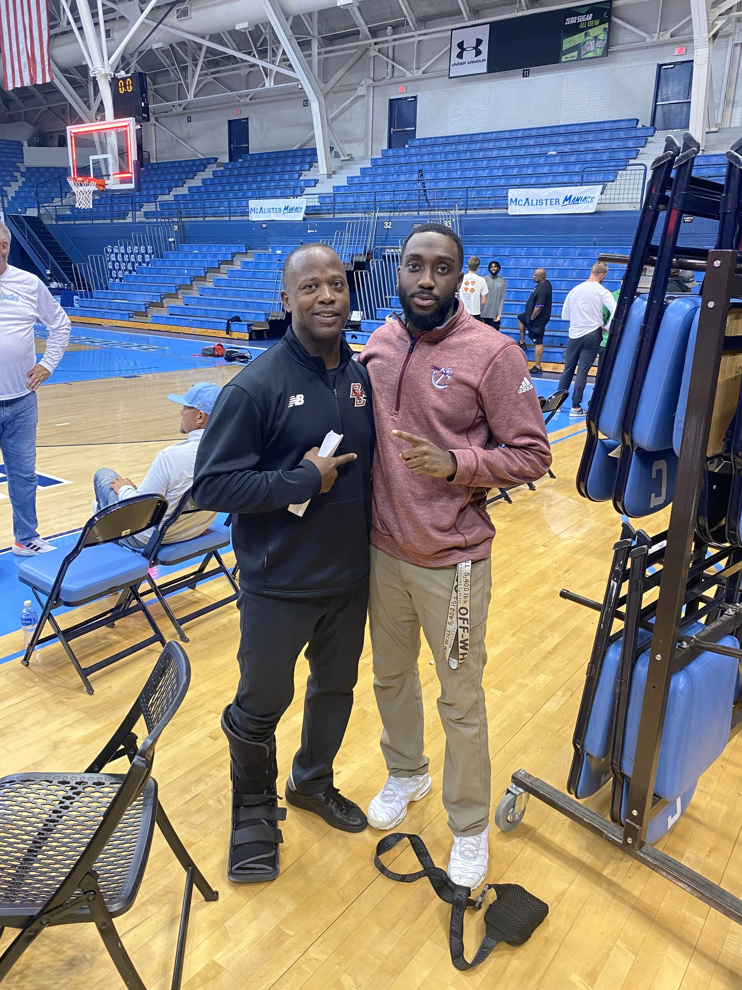 Two men standing on a basketball court inside an arena, posing for the photo. One man is wearing a black sports jacket with a knee brace, and the other is wearing a maroon tracksuit top and khaki pants. There are empty blue bleachers and some chairs 