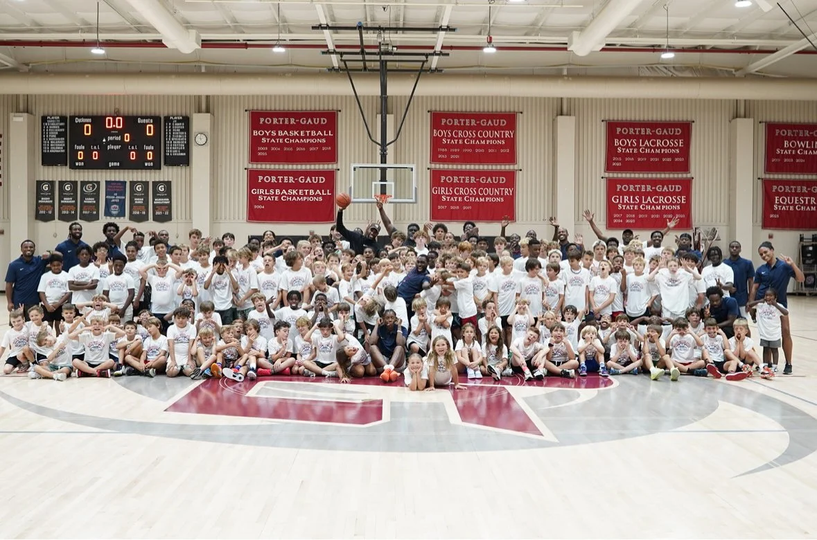 Large group of children and coaches on a basketball court, with many holding basketballs and some making peace signs, in a gymnasium with championship banners.