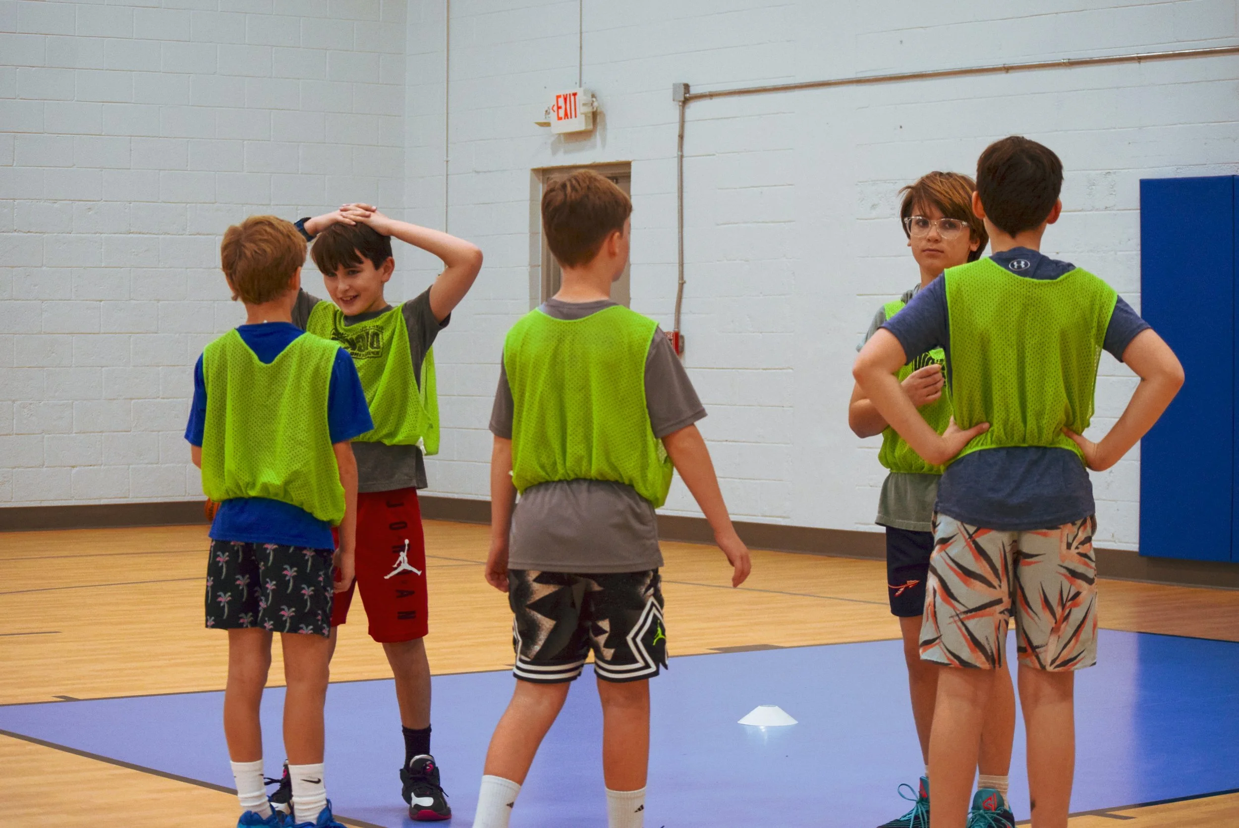 Five children wearing neon green practice vests on a gymnasium floor, engaging in conversation.