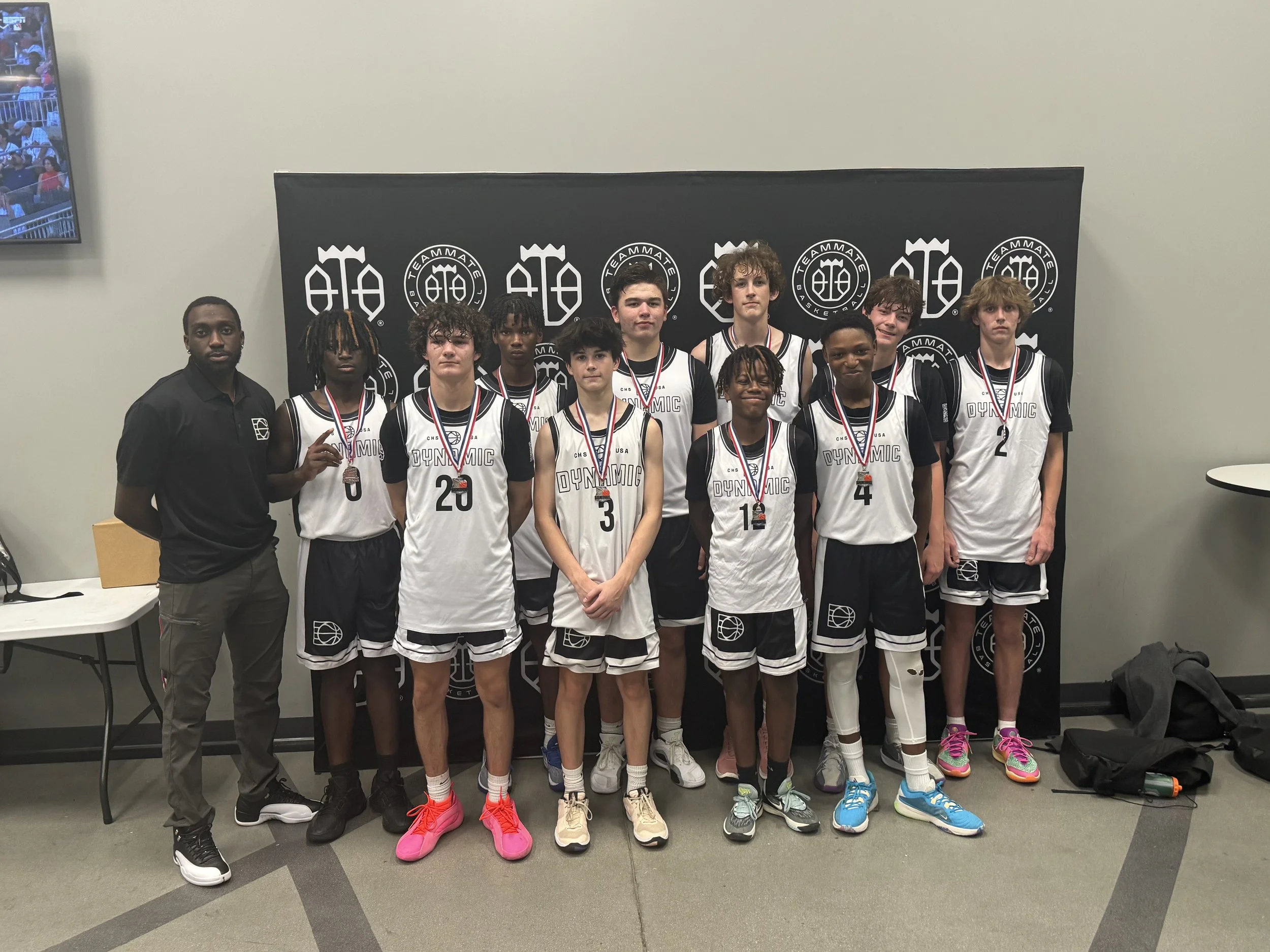 Group of young boys in basketball uniforms holding medals, standing in front of a black backdrop with a sports logo, with their coach on the left side.