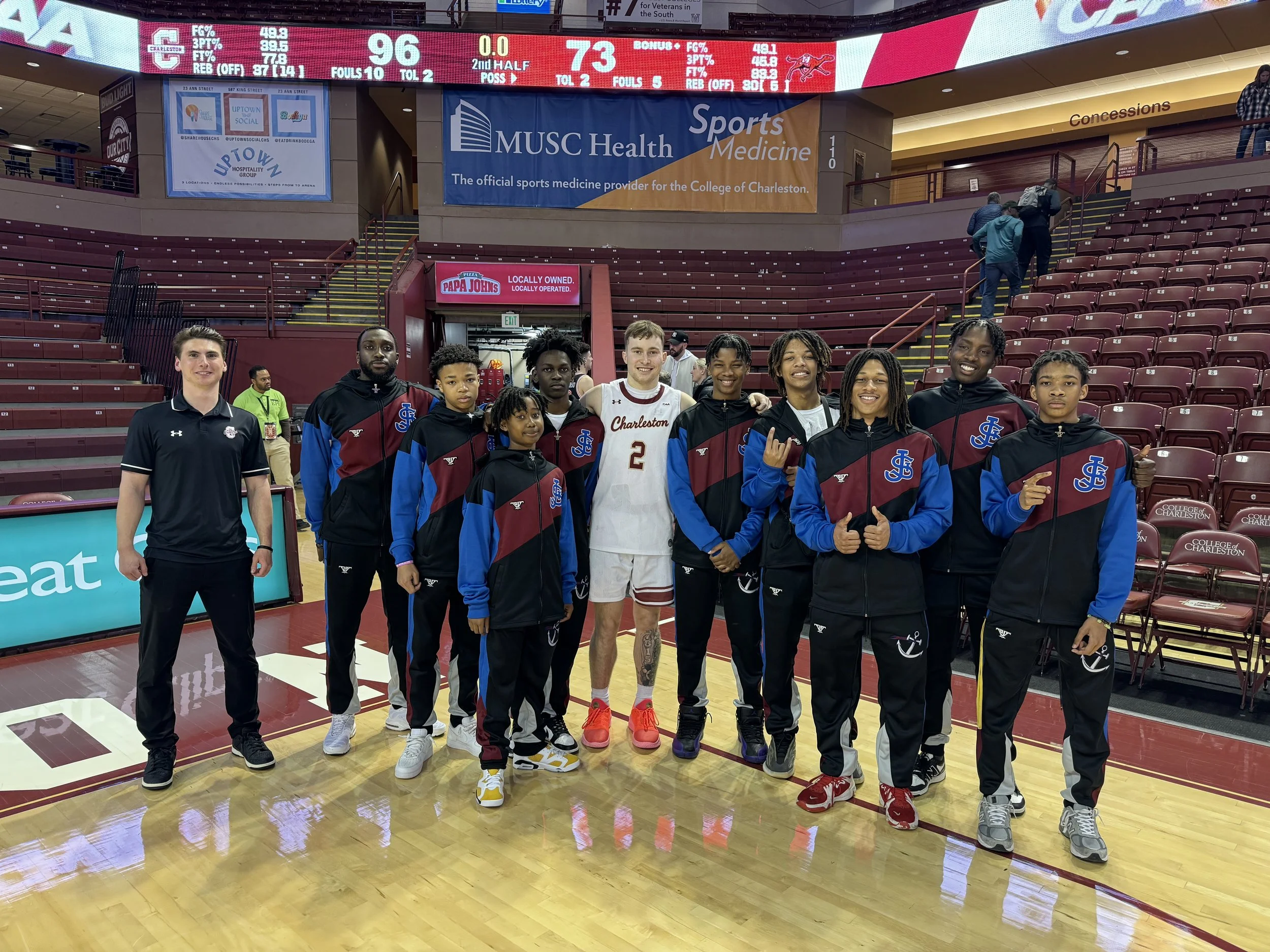 A group of young basketball players and their coach posing on the basketball court after a game, with the score displayed in the background.