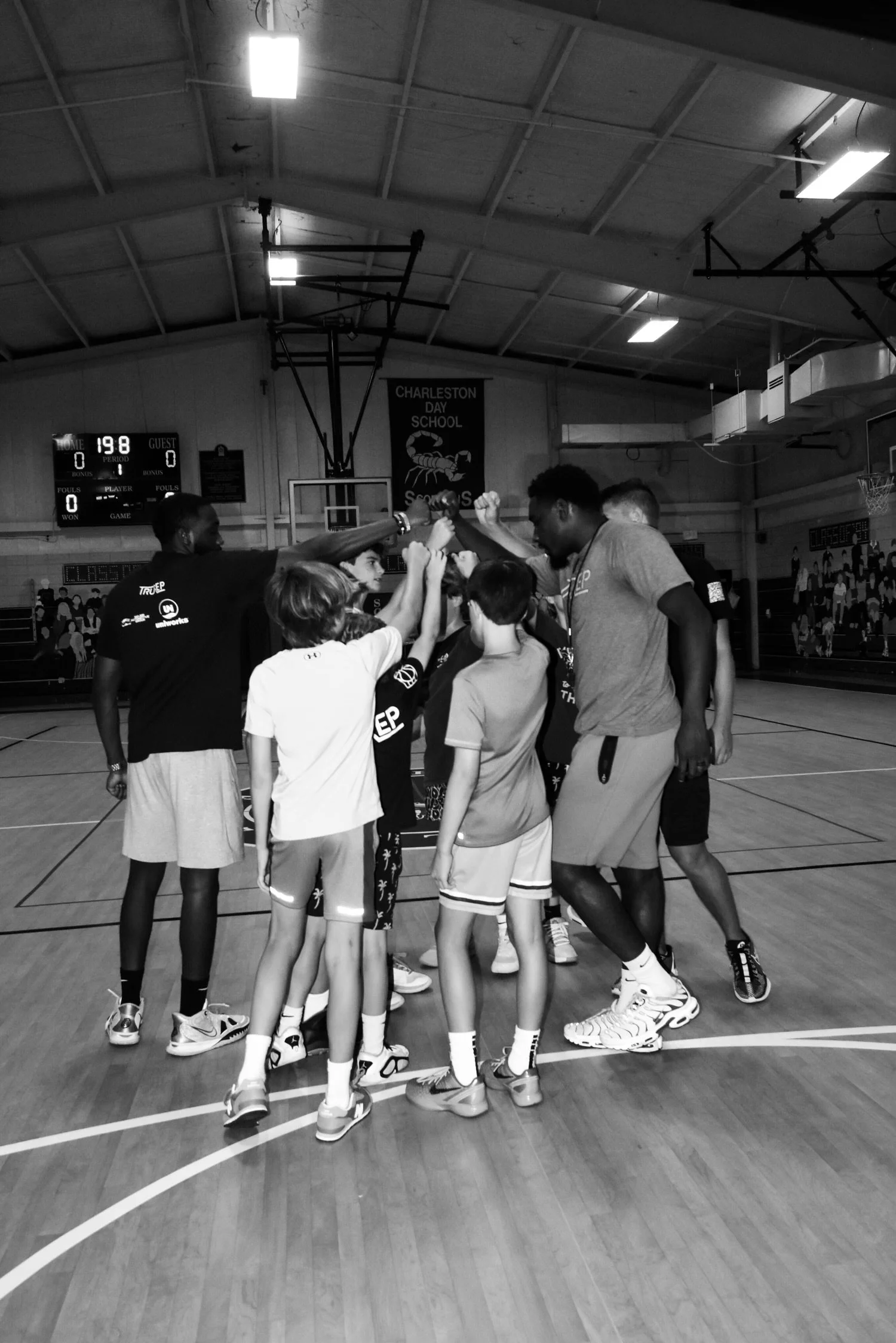 A group of young basketball players and coaches huddle together in a gym, raising their hands in a team cheer.