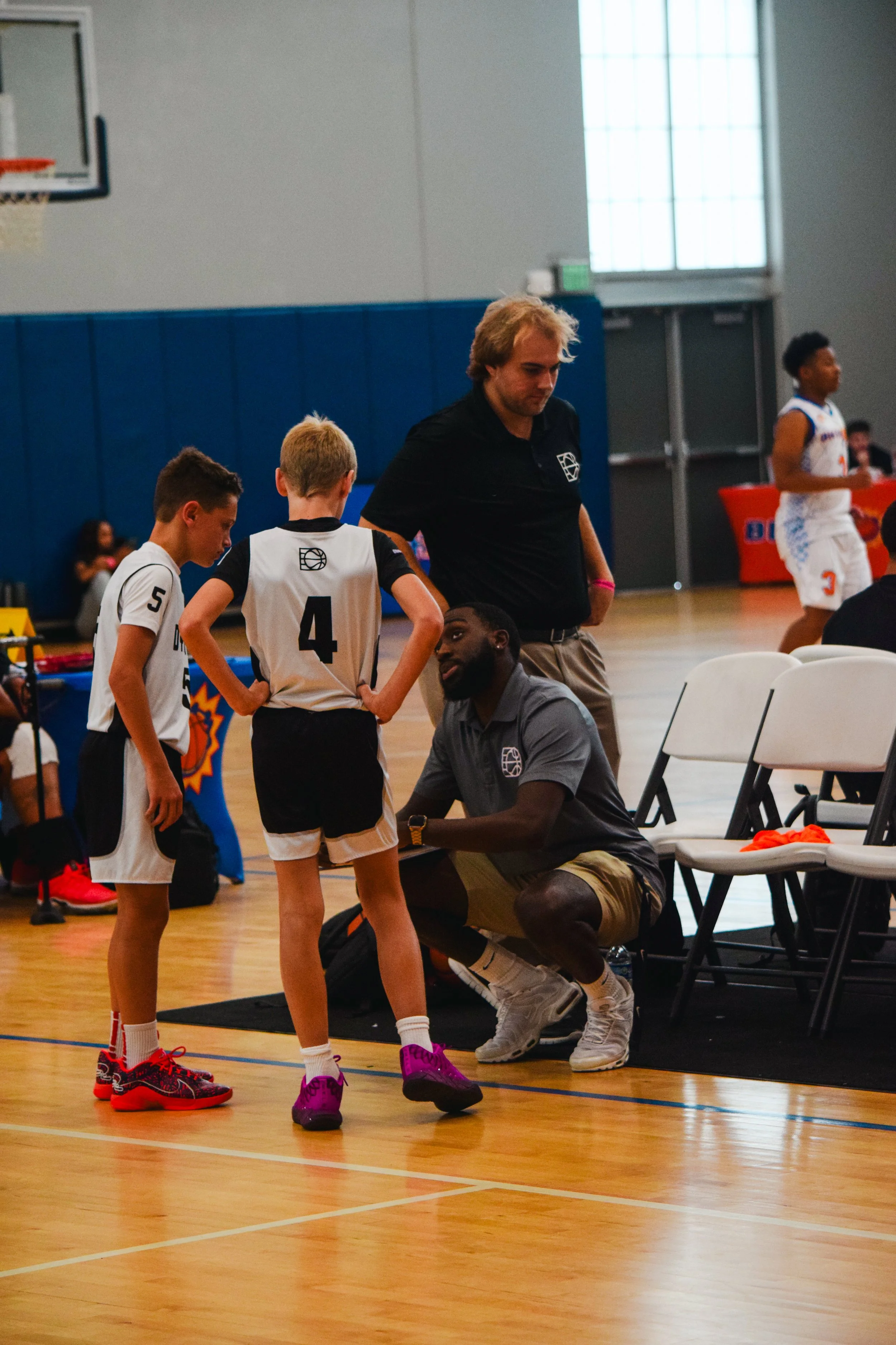 A basketball coach talking to two young players during a game in an indoor gym, with a player in the background.