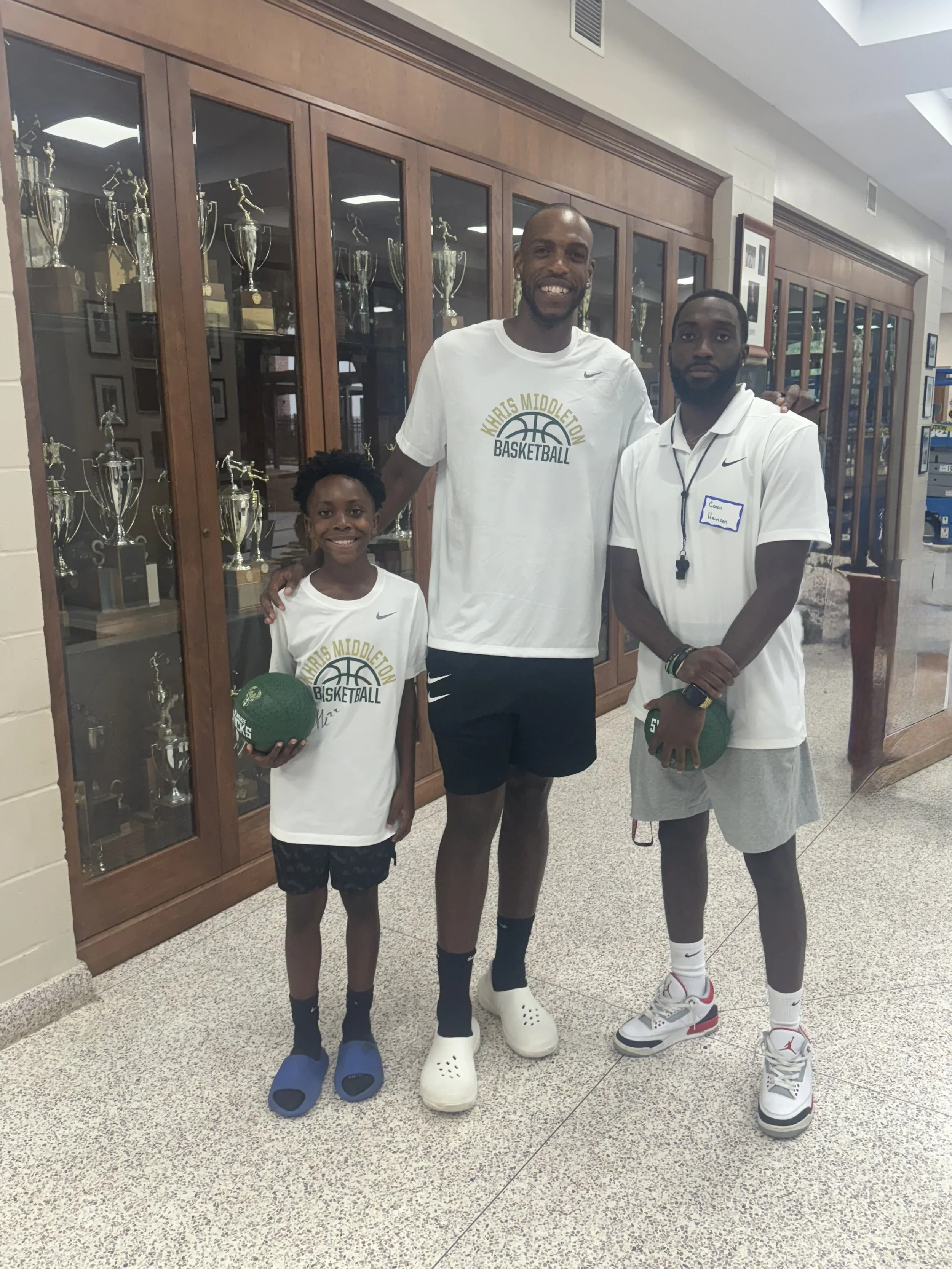 Three individuals posing in front of a trophies display at a school. All are wearing white T-shirts with 'Karis Middleton Basketball' logos. The young girl on the left is holding a green basketball with the word 'SPECKS' on it, wearing black shorts a