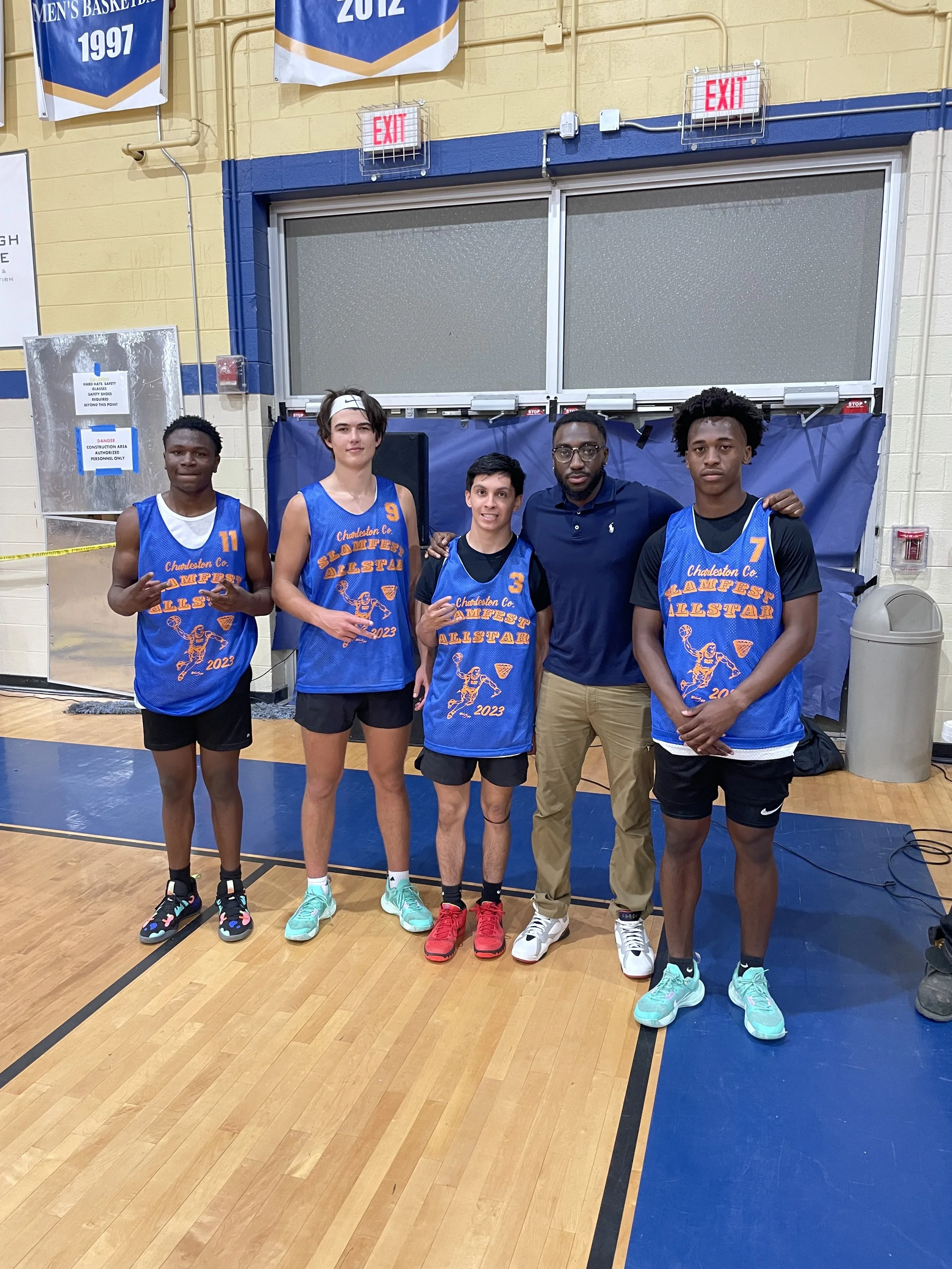 Group of five people standing in a gymnasium, four young male basketball players in blue jerseys with orange and white lettering, and one adult man in casual clothing. The players are posing with their coach after a basketball game or event, with ban