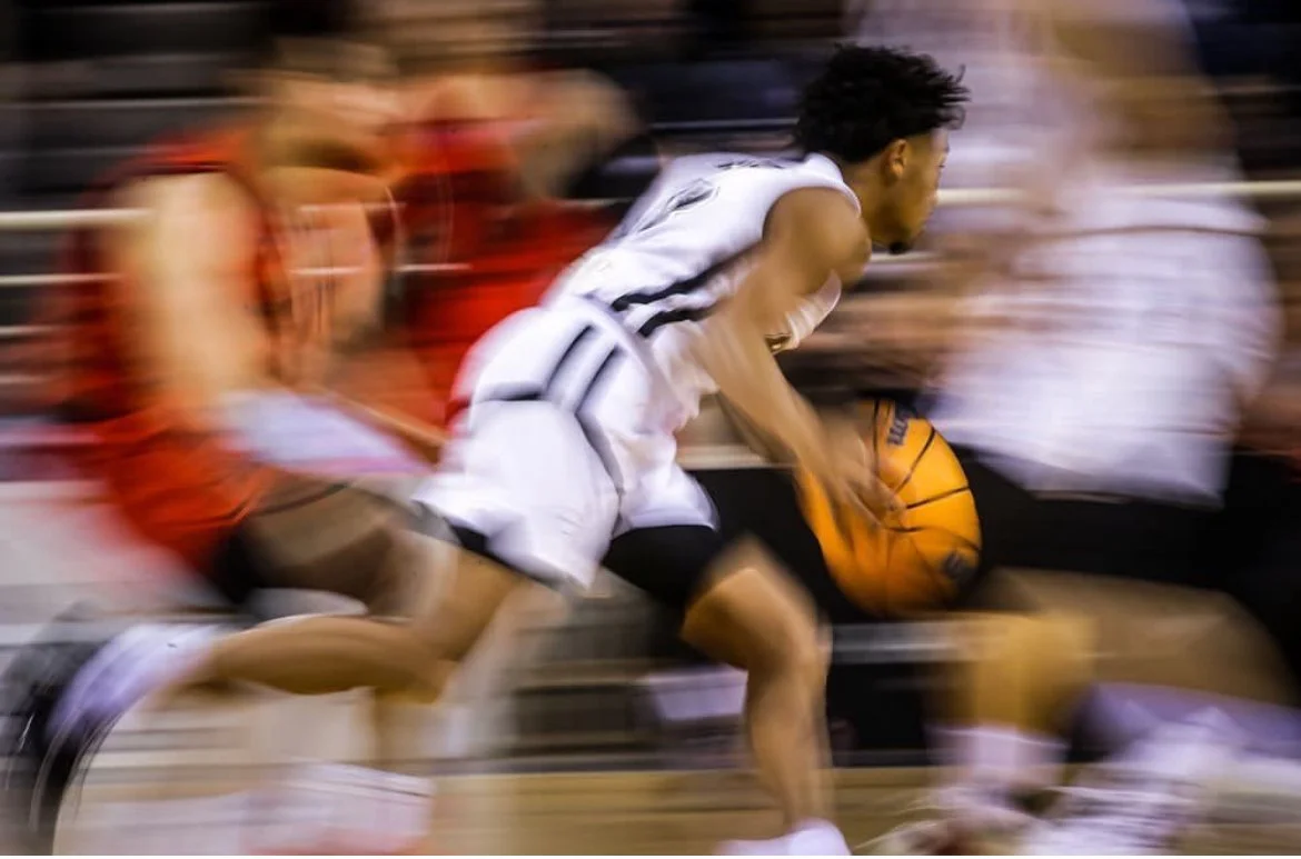 A basketball player in white uniform dribbling the ball, blurred motion indicating fast movement during a game.