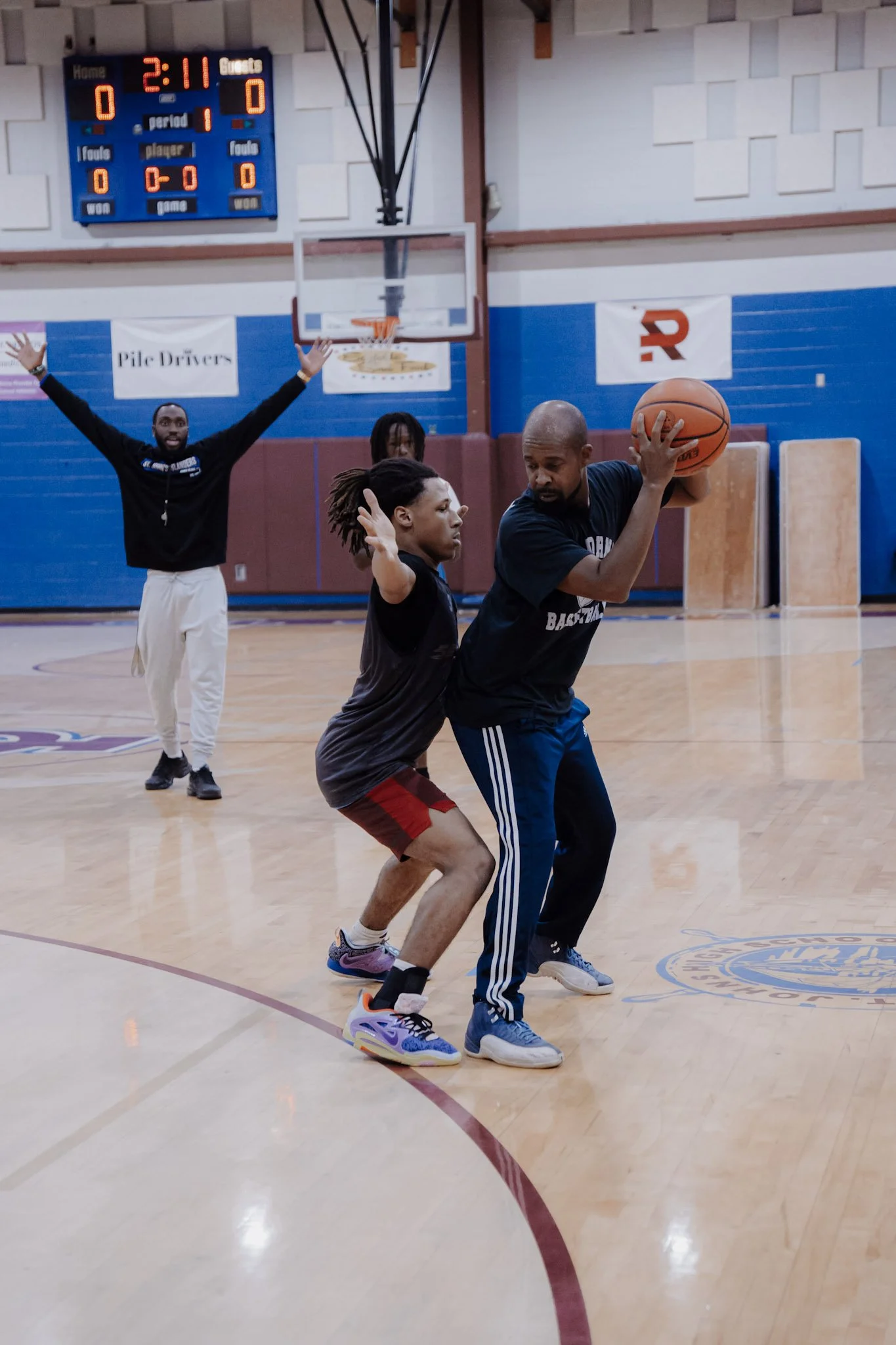 Three men playing basketball in an indoor court; one is holding the ball, another is guarding closely, and the third is in the background with arms raised.