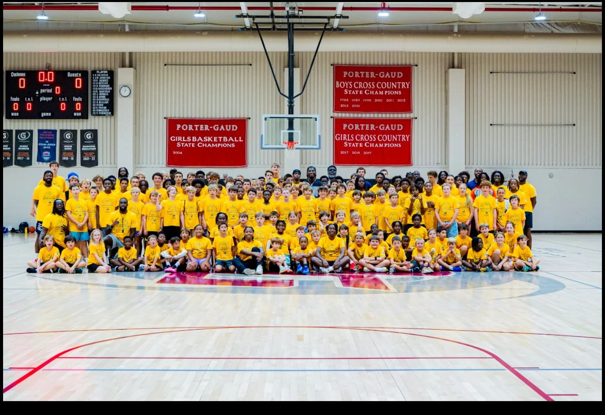 A large group of young children and a few adults in yellow T-shirts posing for a photo in a basketball gymnasium with banners on the wall.