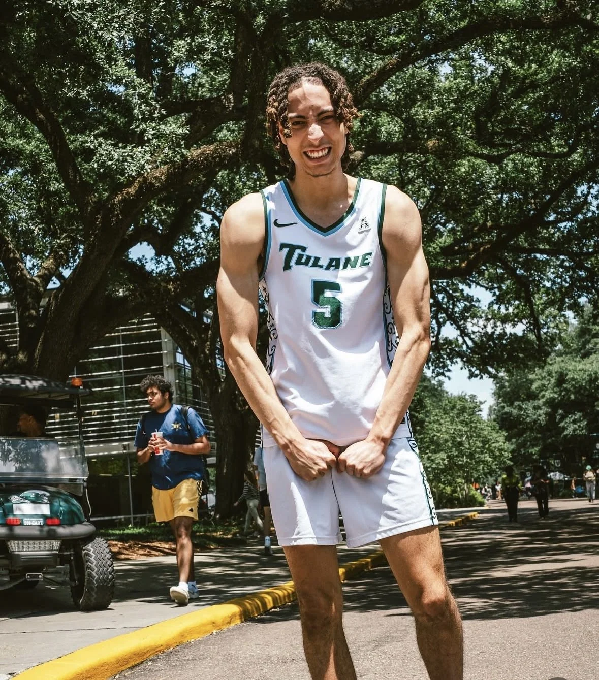 A young man in a white Loyola University basketball jersey with the number 5, standing outdoors under a large tree, smiling and clenching his fists.