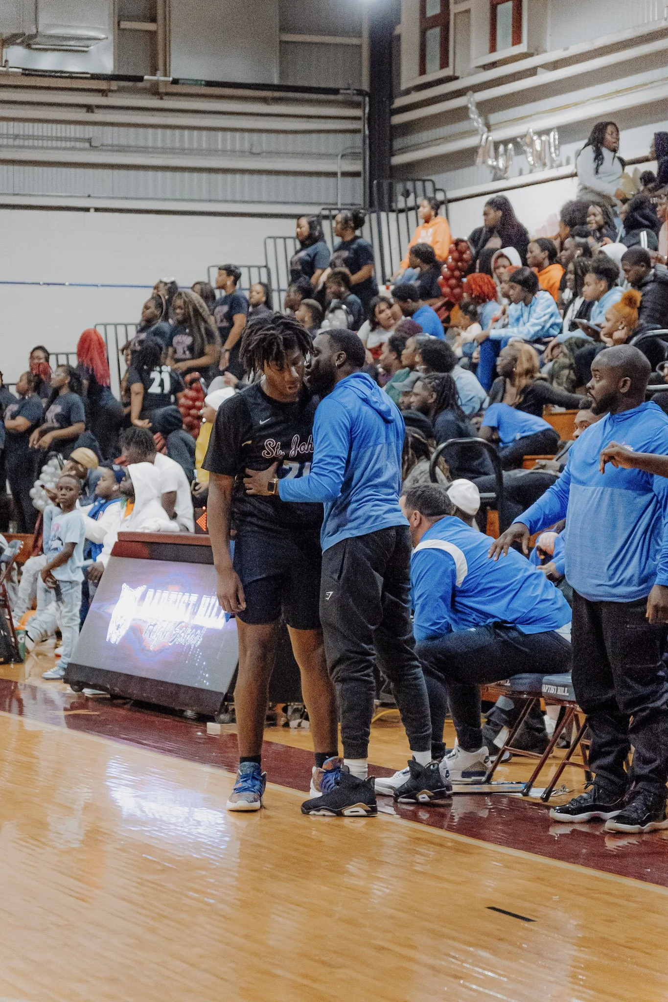 A basketball player in a black uniform with 'St. John' on it is talking to a coach wearing a blue jacket, amidst a crowd in a gymnasium.