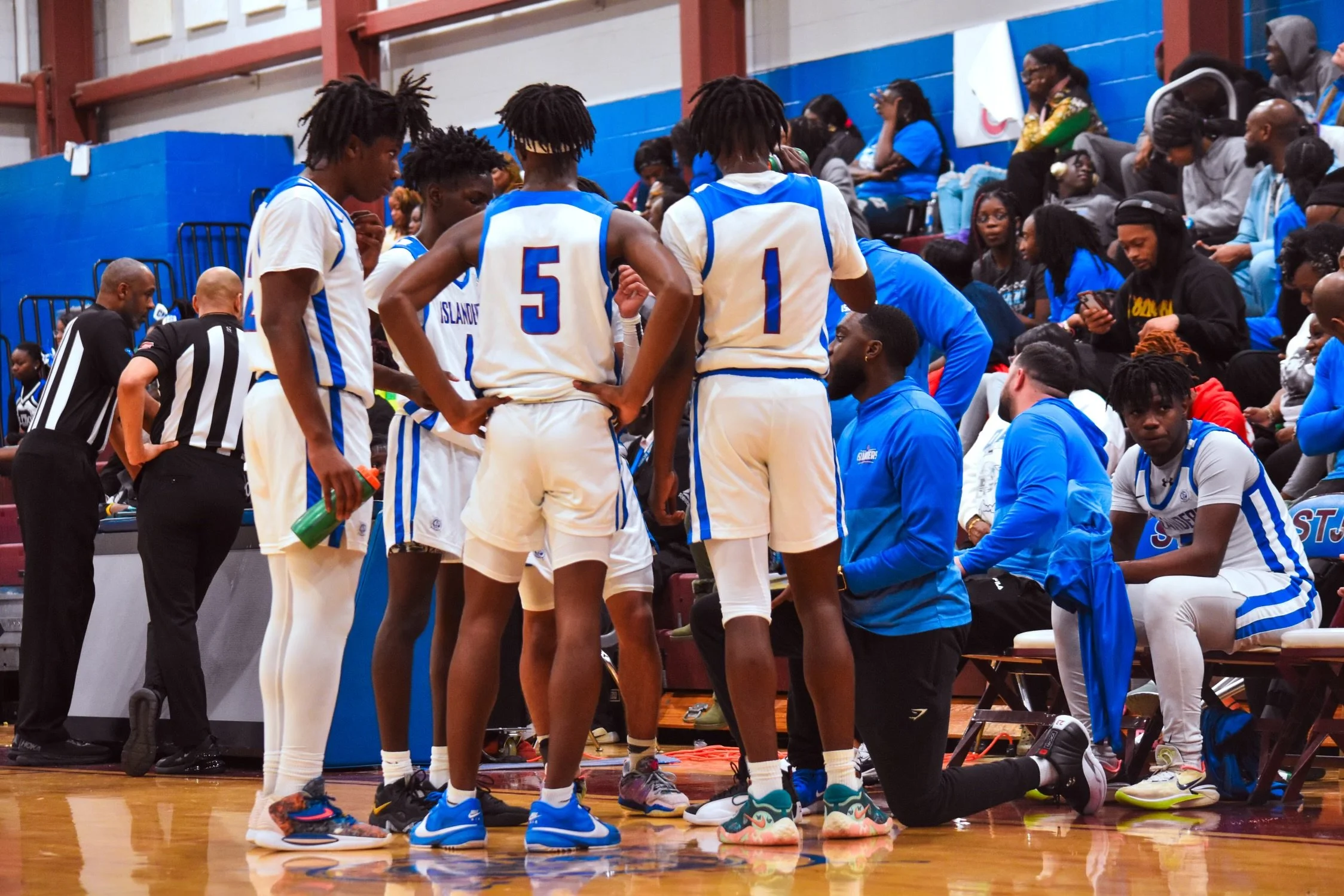Basketball team in white and blue uniforms huddles around their coach during a game or timeout, with spectators seated in the background.
