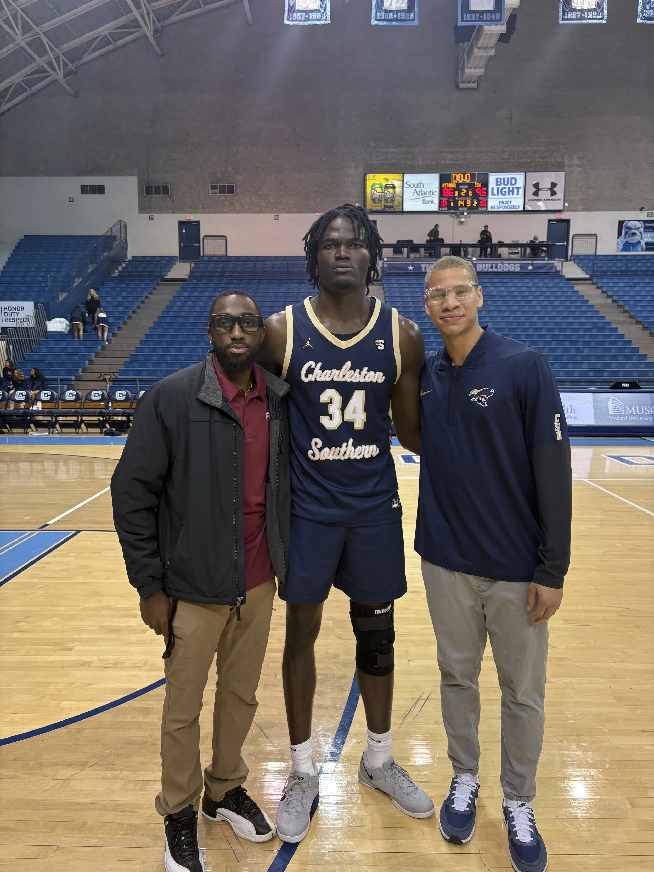 Three men standing on a basketball court, one in the middle wearing a navy Charleston Southern basketball jersey with the number 34, and the two men on either side in casual clothing, inside a sports arena with banners hanging from the ceiling and em