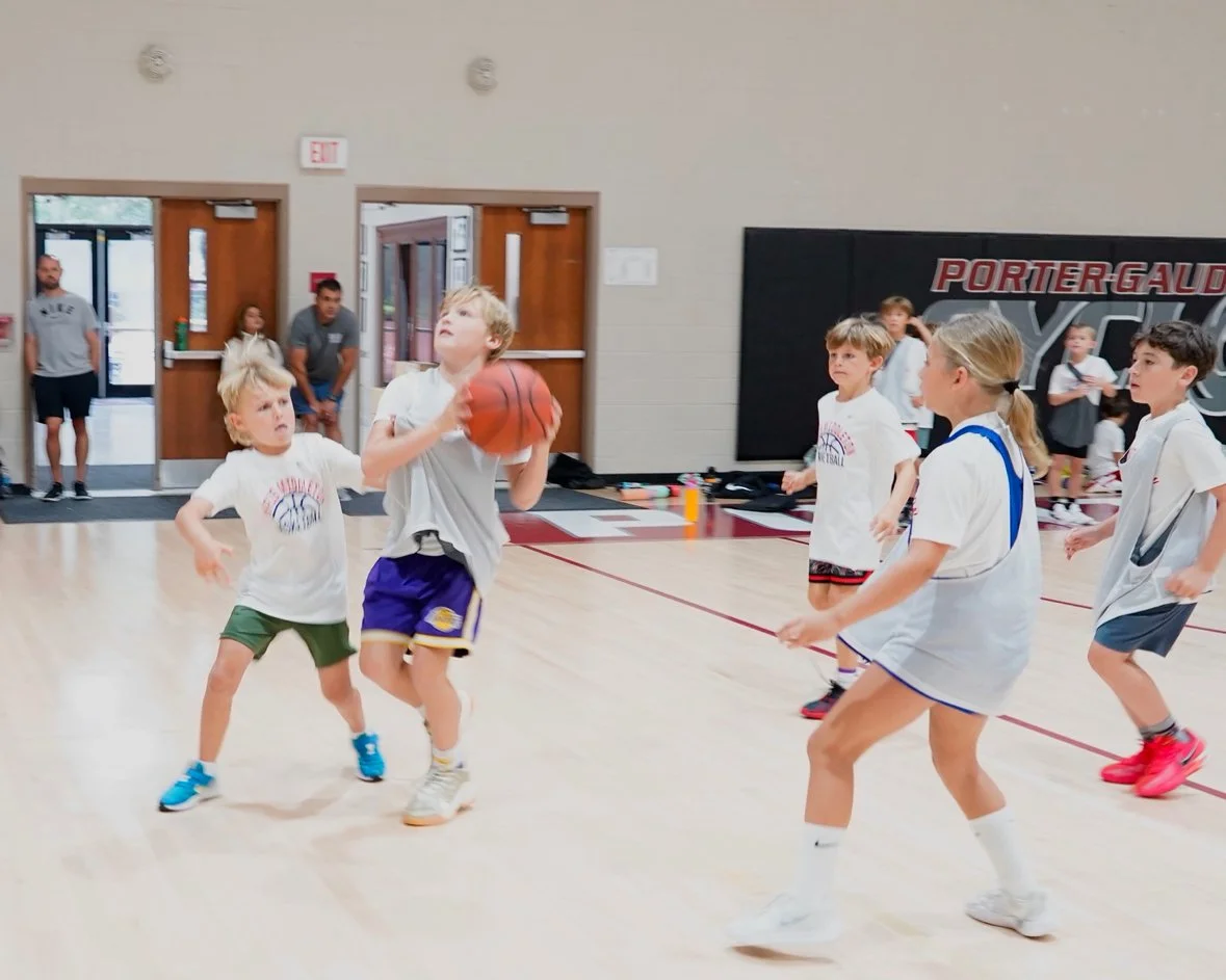 Young children playing basketball in an indoor gym. One boy is holding the basketball, and others are watching or preparing to play.