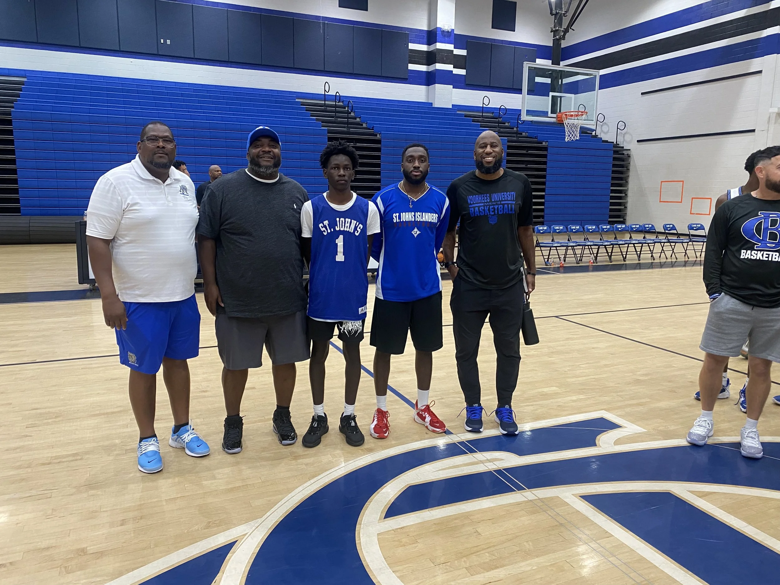 Group of five men standing on a basketball court with blue and white seating in the background. Some are wearing basketball jerseys and casual clothing.
