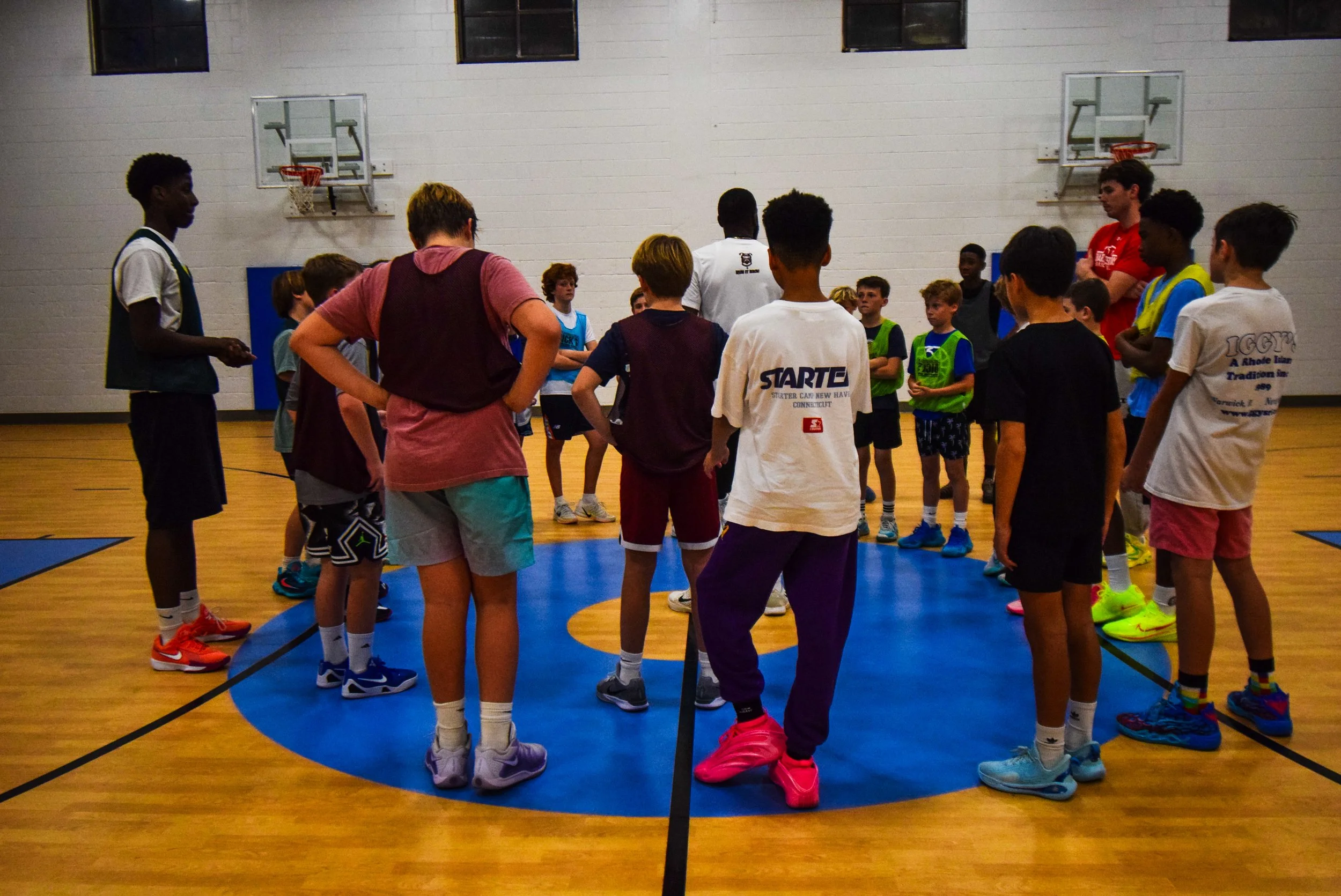 A group of children and a coach gathered in a circle inside a gymnasium for a basketball practice or team meeting.