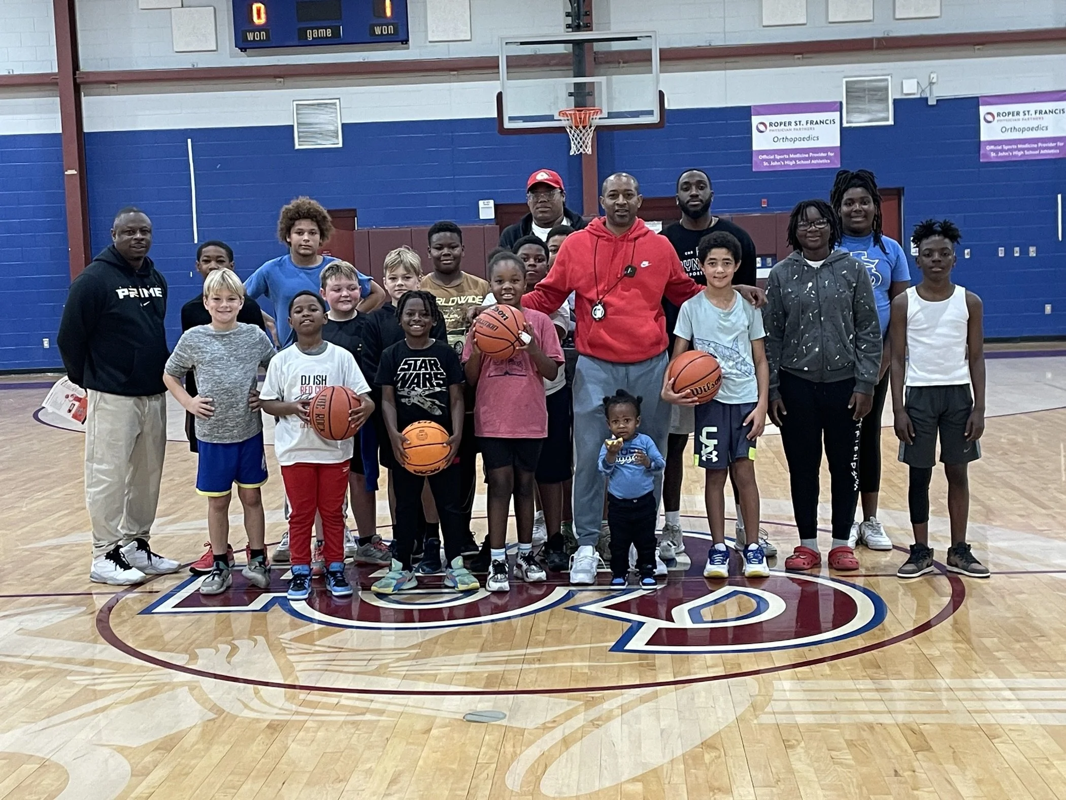 A group of children and adults posing on a basketball court inside a gymnasium, with some holding basketballs.