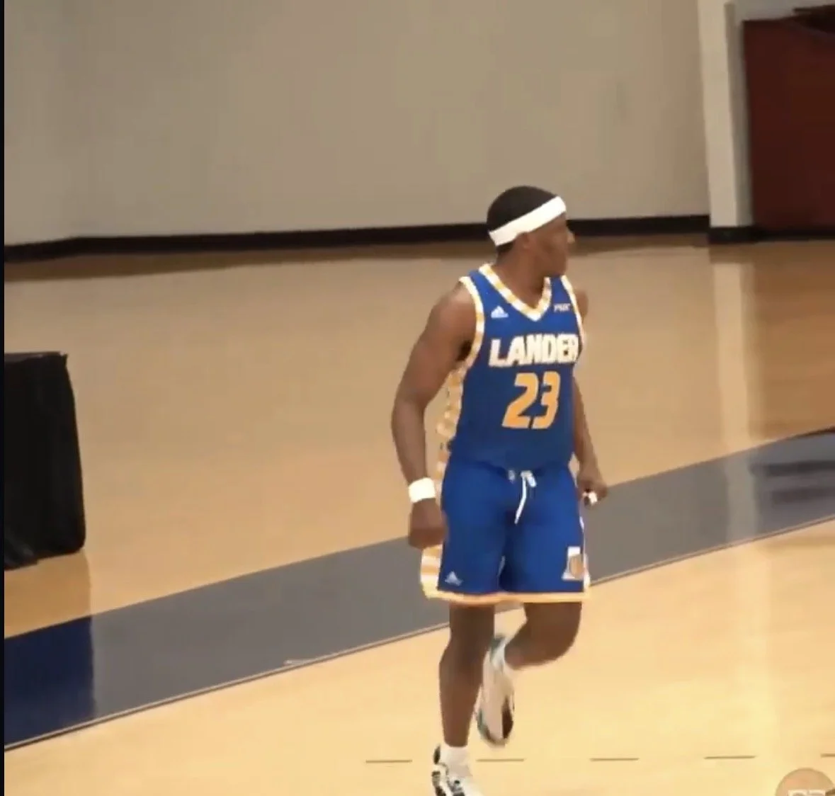 A male basketball player in a blue and yellow uniform holding a dumbbell in his right hand, standing on a basketball court, wearing a white headband and white wristband, with the number 23 on his jersey.