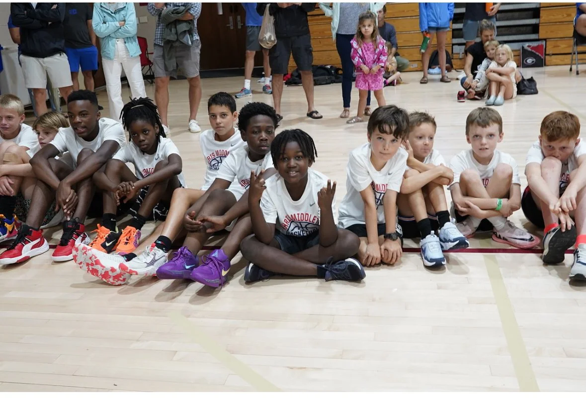 Young boys and girls sitting on a gymnasium floor, some in basketball uniforms, with more children and adults standing or sitting in the background.