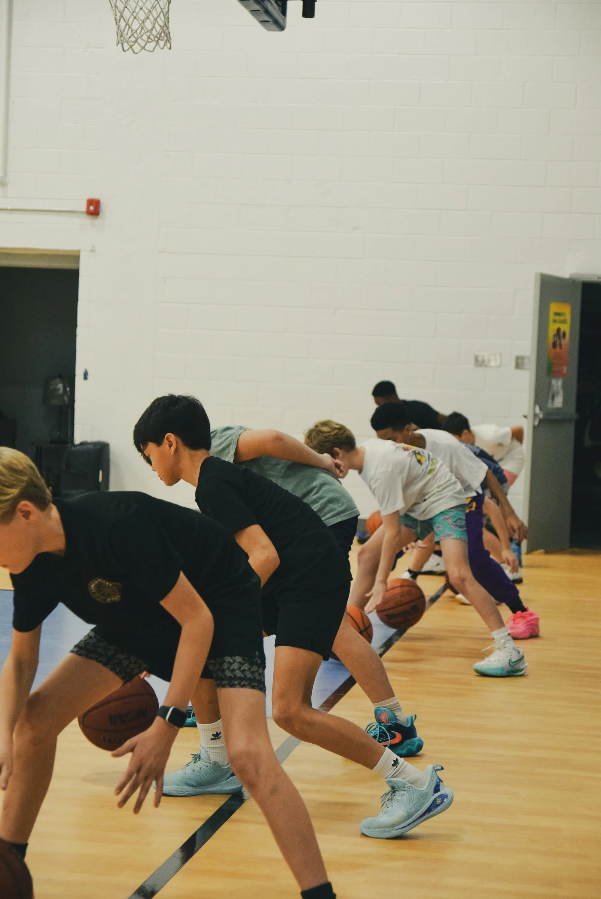 Children practicing basketball dribbling in a gym, lined up along a wall, with basketballs in hand.