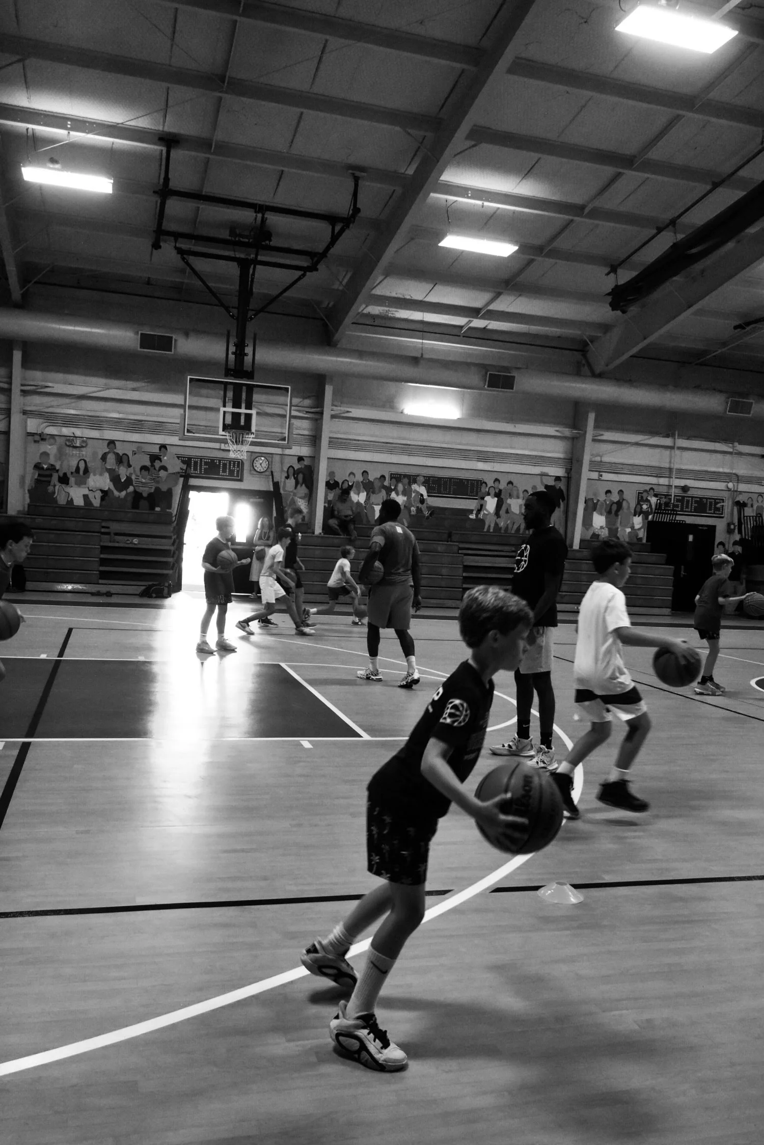 Children participating in a basketball practice session in a gymnasium with a mural of spectators on the wall.